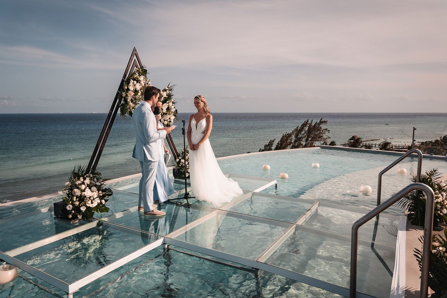 Wedding ceremony on glass-floored oceanfront deck with triangular floral arch.
