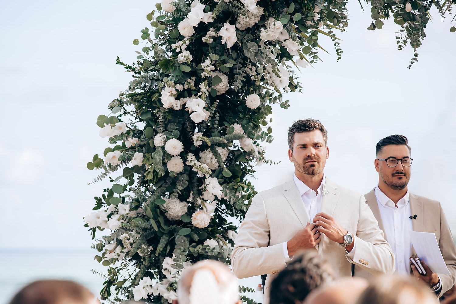 A wedding ceremony takes place under a floral arch with white roses and greenery in an outdoor garden setting.