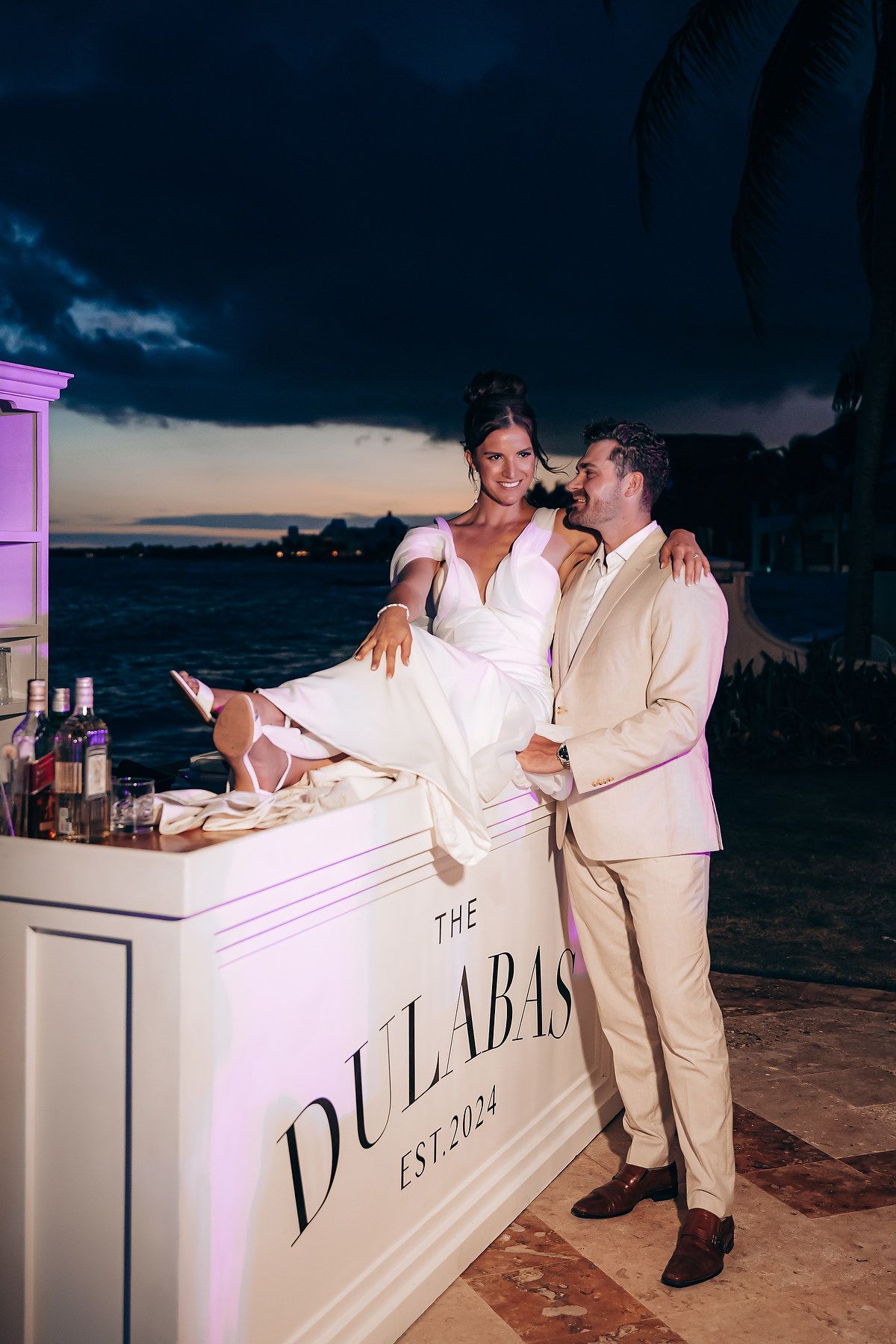 Couple in white outfits sharing intimate moments at an illuminated beach bar during sunset.