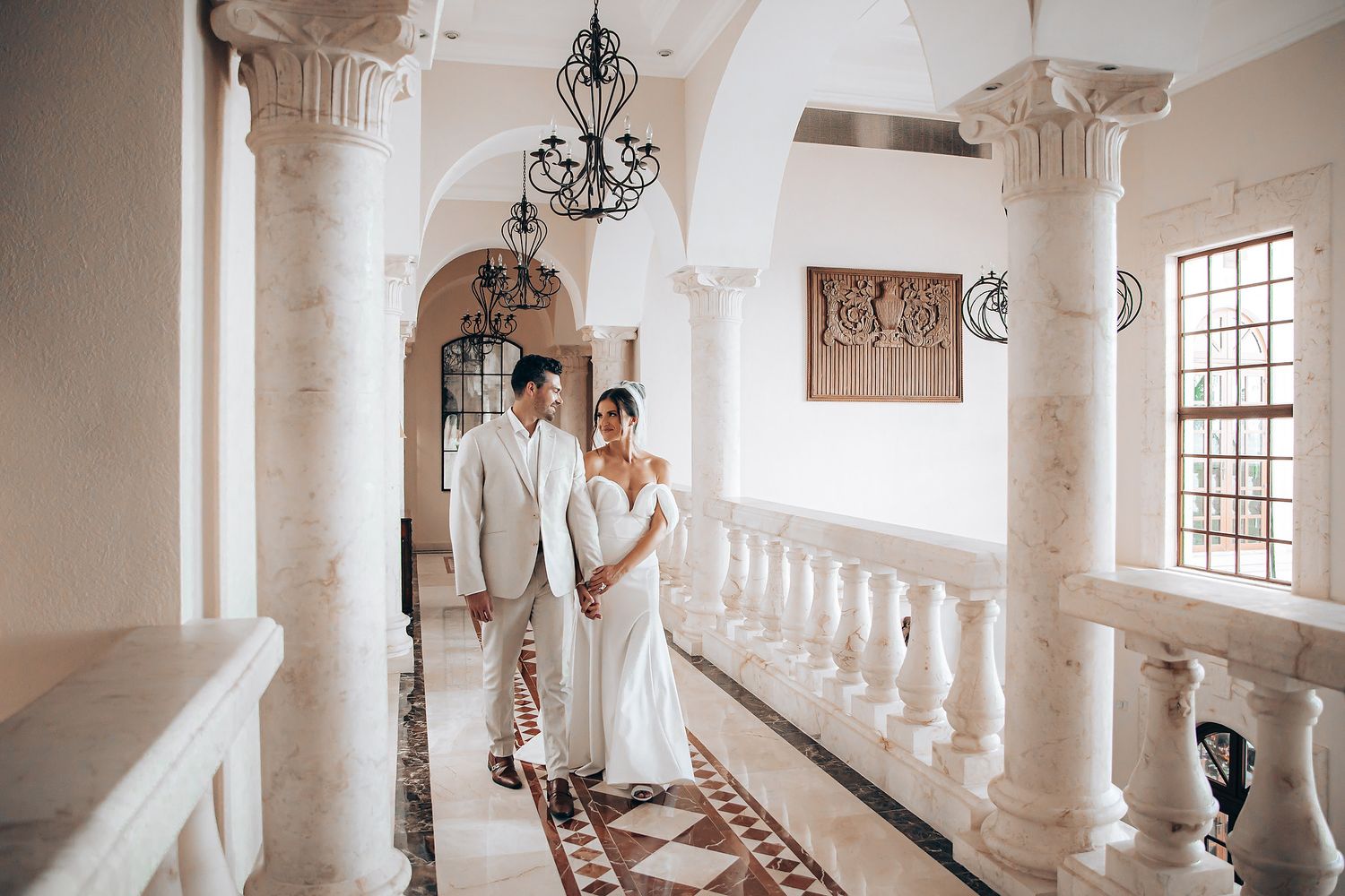 A romantic couple in wedding attire poses in a bright white hallway with ornate columns and arched windows.