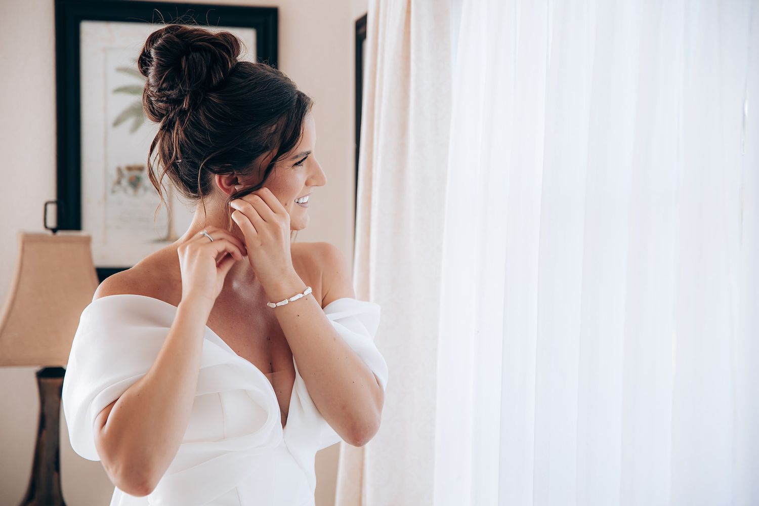 A bride in a white dress adjusts her earrings while getting ready in front of a mirror.