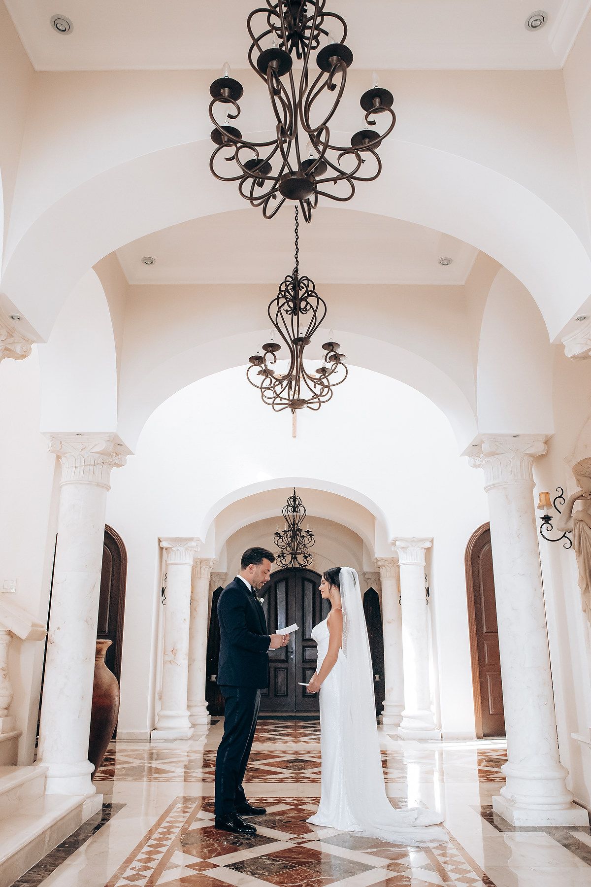 Elegant white colonnade hallway with ornate black chandeliers and arched ceilings in a wedding venue.