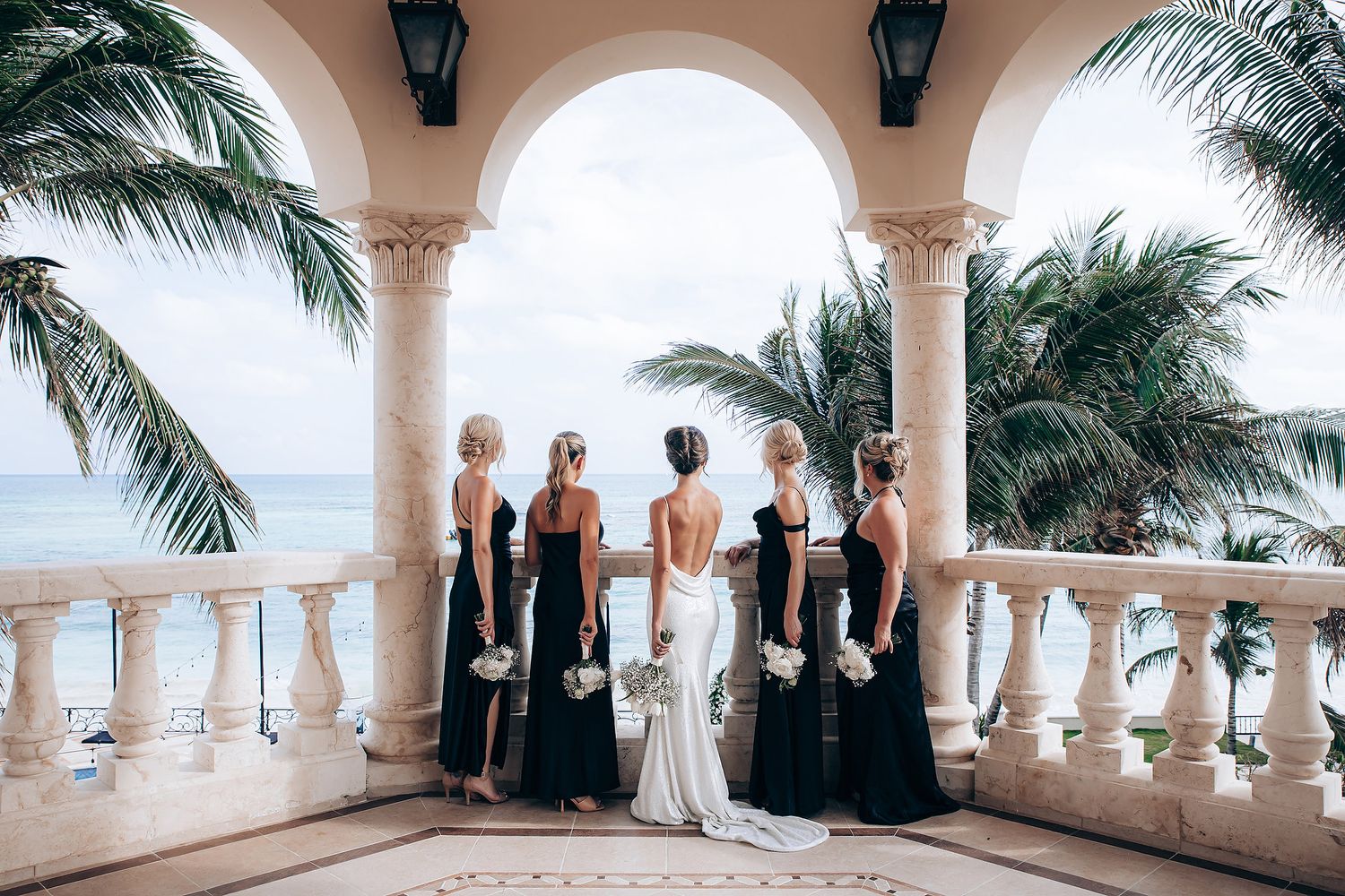 Wedding party gathers under elegant white arches overlooking a tropical beach and ocean view.