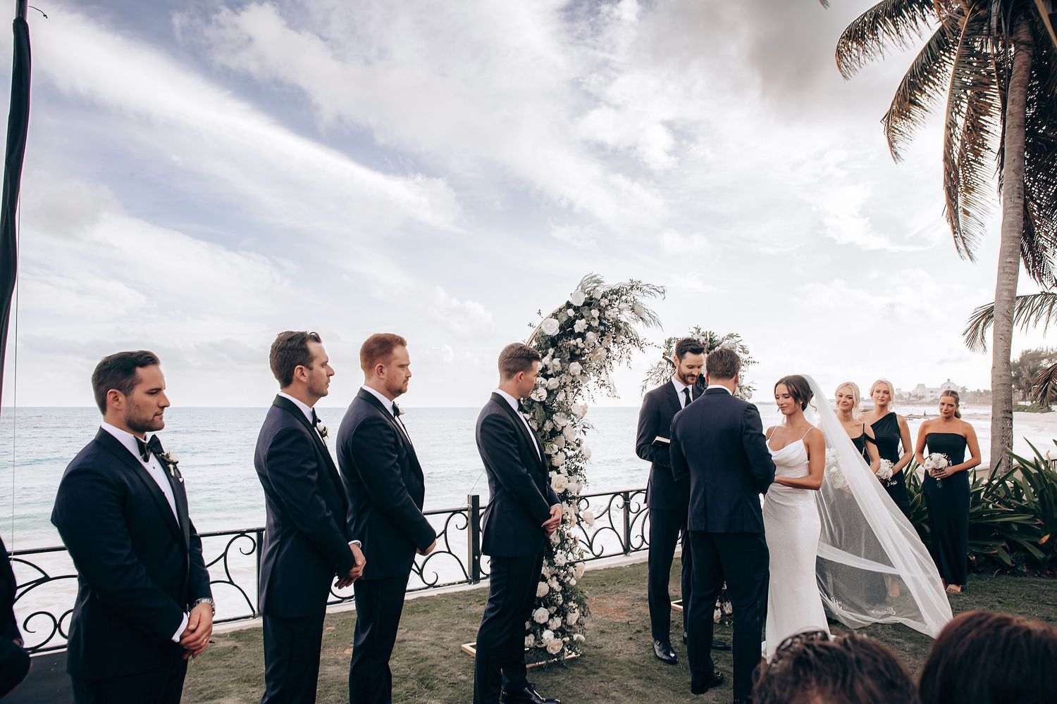 A wedding ceremony takes place on an oceanfront terrace with palm trees and tropical scenery in the background.