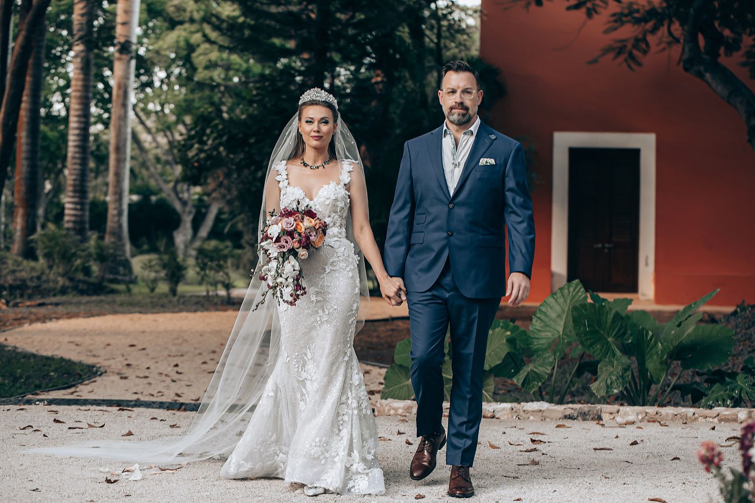A couple in wedding attire poses for romantic photos on a path near a red barn in a rustic outdoor setting.
