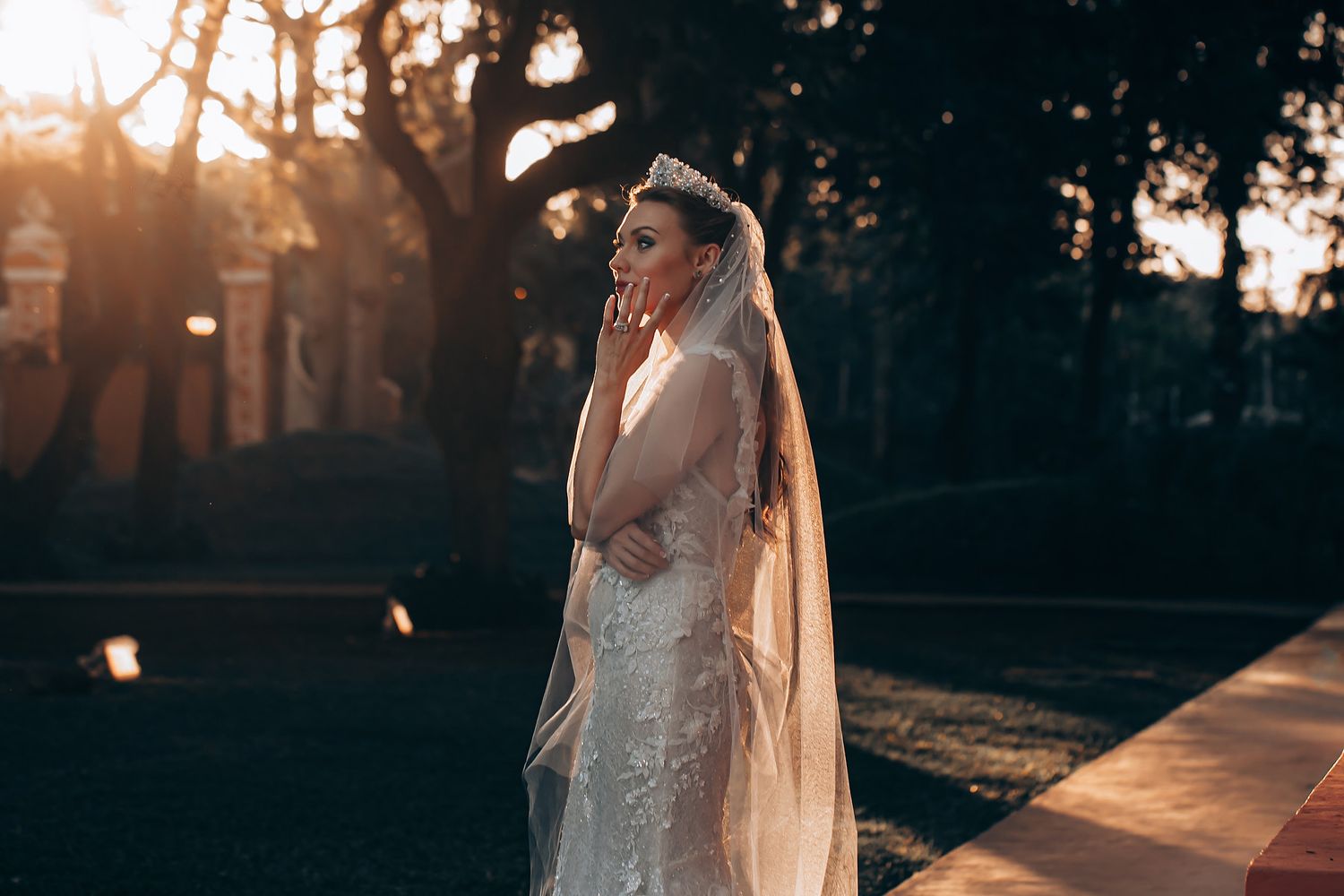 A bride in a lace wedding dress poses in golden sunlight during sunset at an outdoor venue.