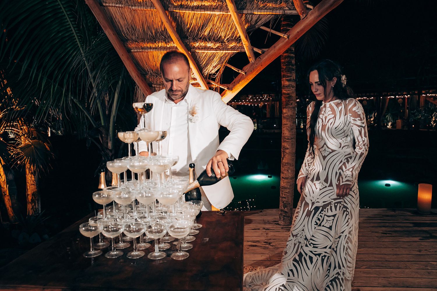 A person in white pouring champagne into a tower of glasses at a tropical evening event while another guest watches.