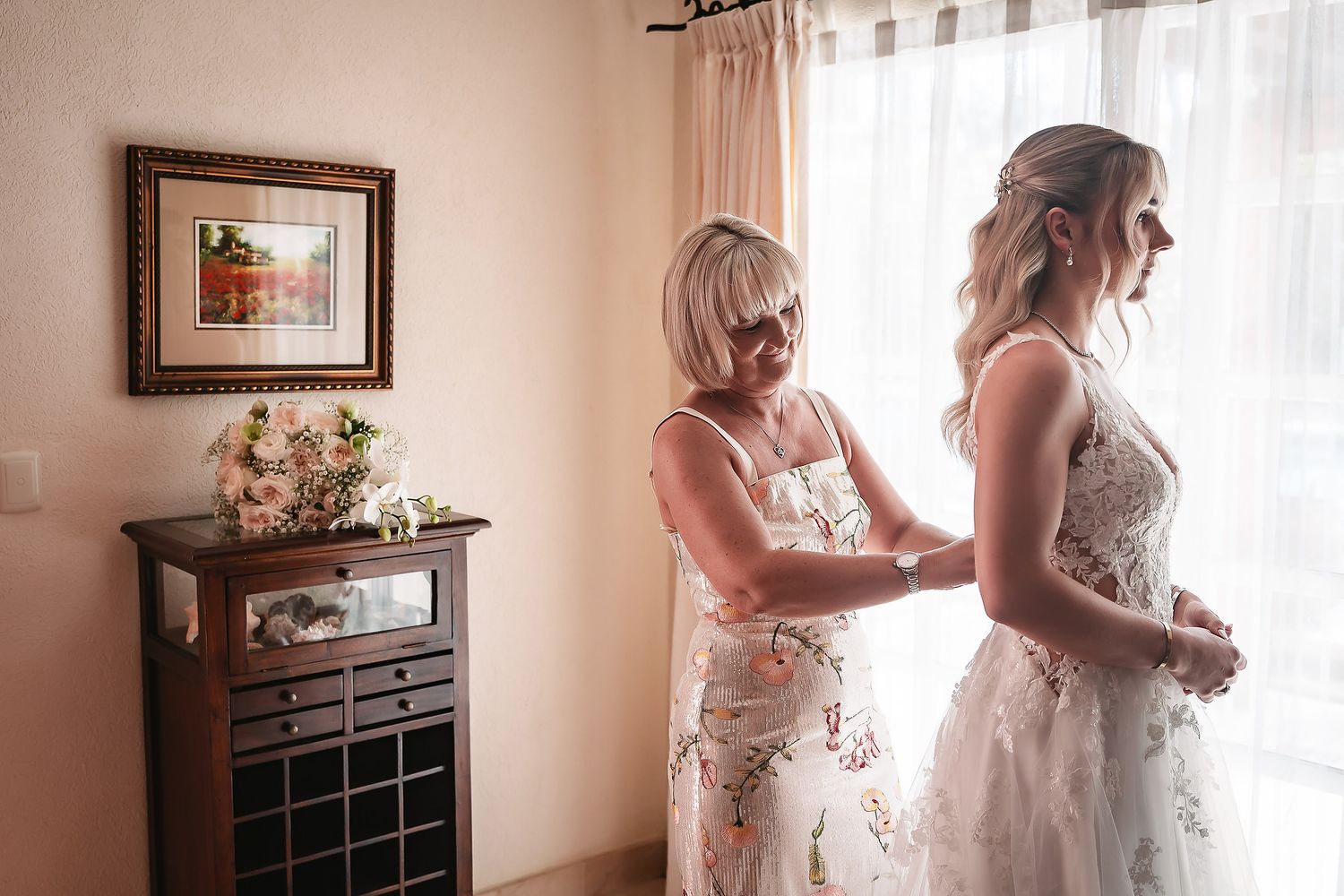 Women in floral dresses help with wedding preparations in a sunlit room.