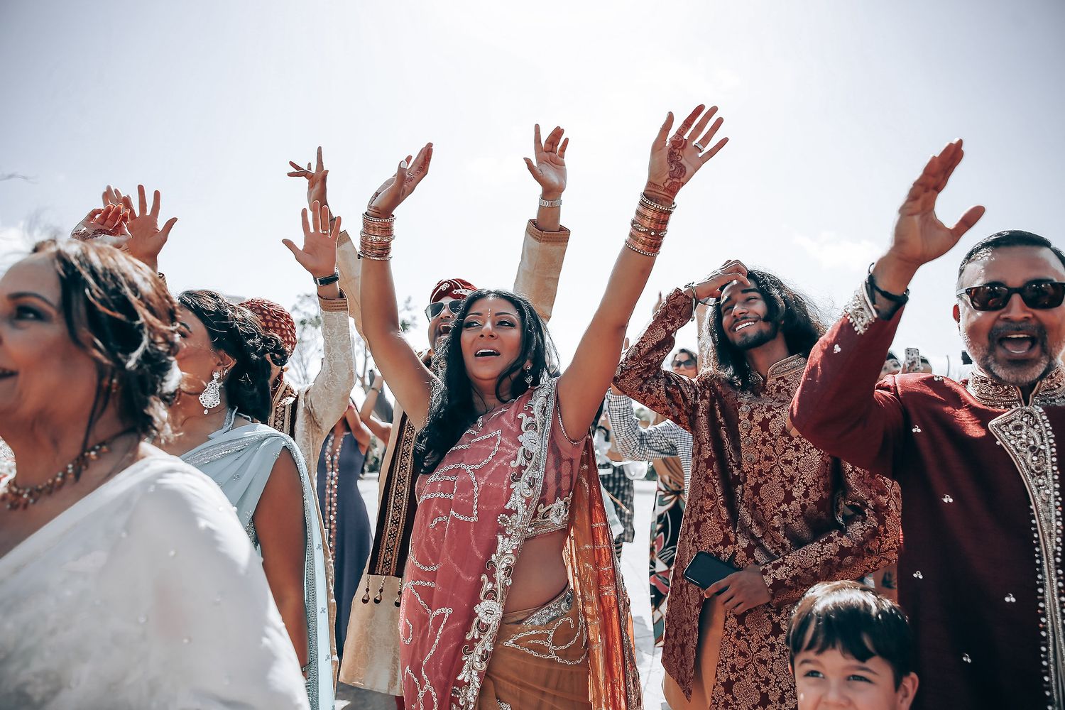 Wedding guests in colorful traditional Indian attire dancing with raised hands at a celebration.