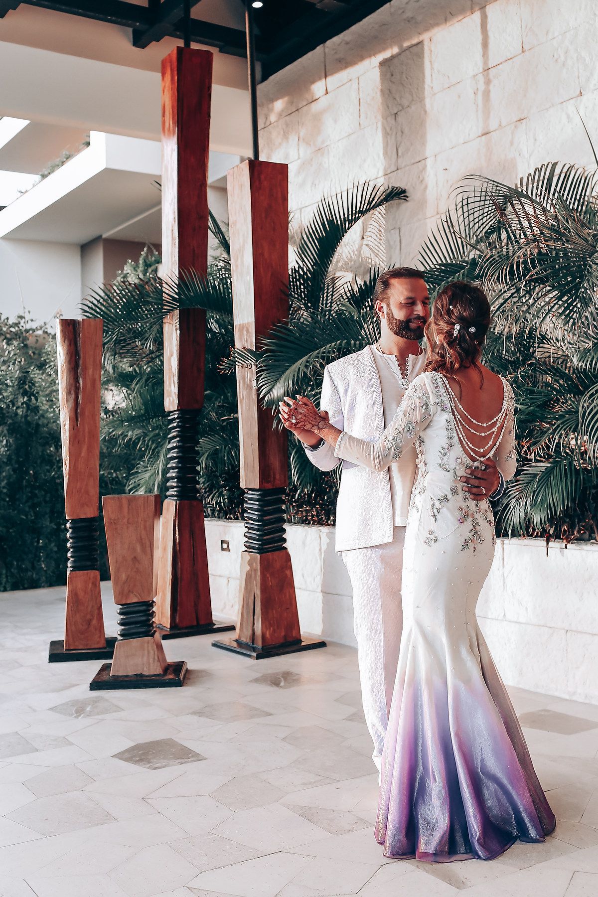 A couple in white and purple ombre wedding attire dance together on a covered patio with palm trees in the background.
