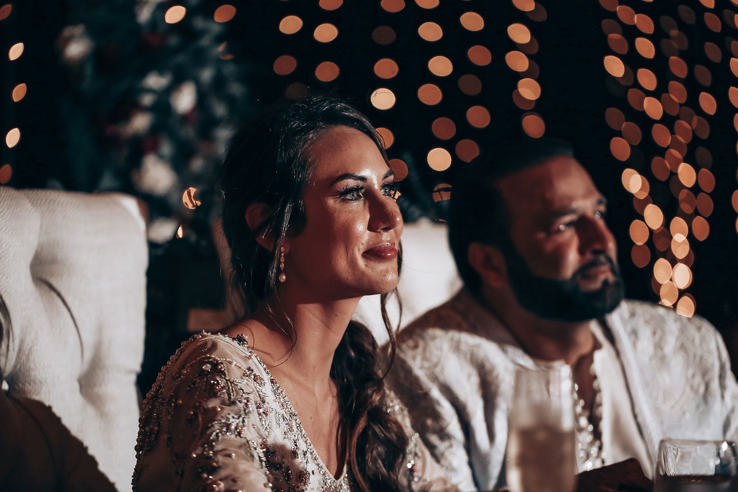 A couple sits together at their wedding reception with twinkling lights in the background creating a romantic ambiance.