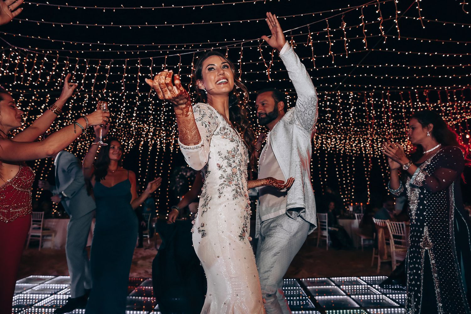 Wedding guests dance and celebrate under twinkling string lights during an outdoor evening reception.