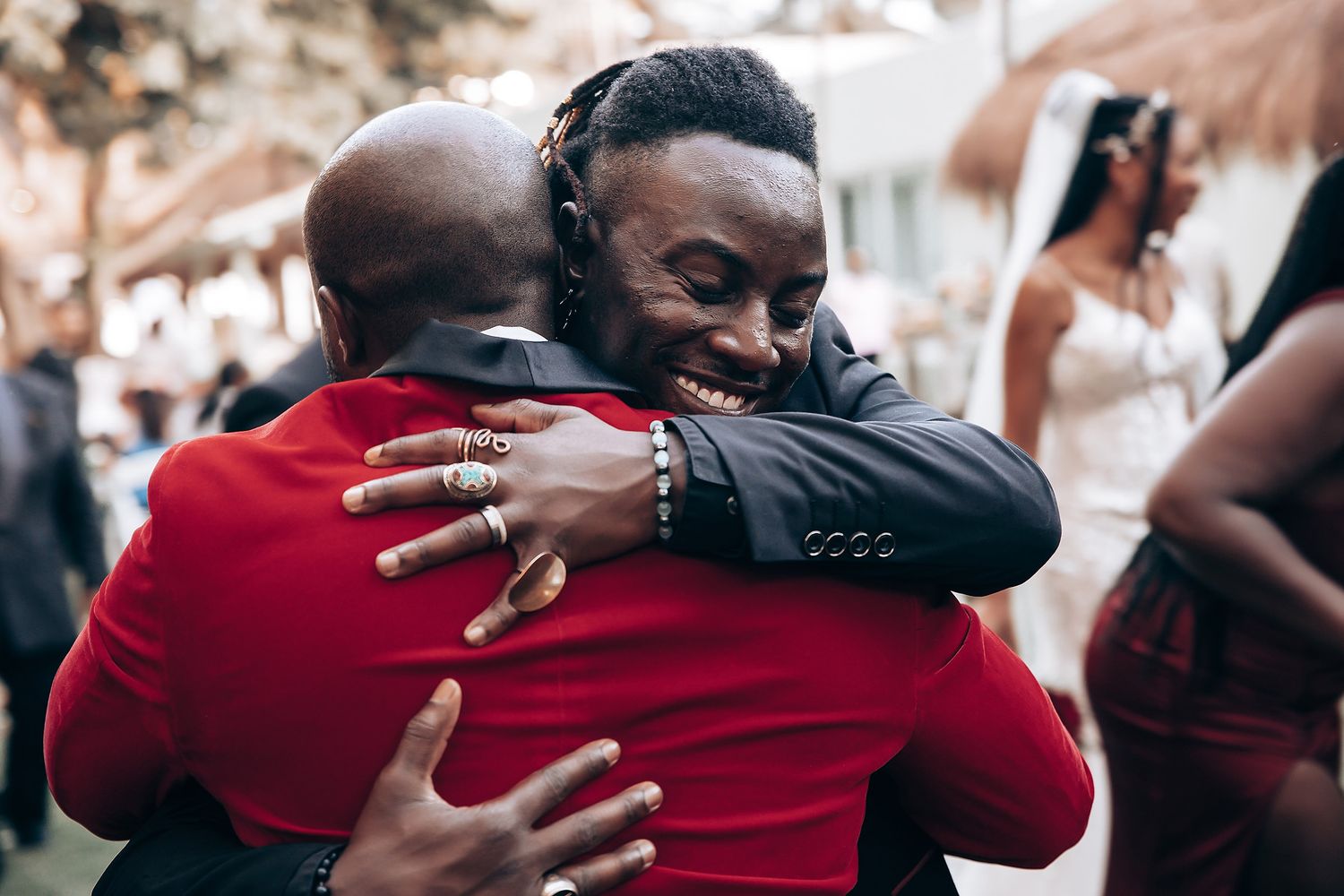 Two people share a warm embrace at an outdoor gathering, one wearing a red shirt and the other in a dark suit.