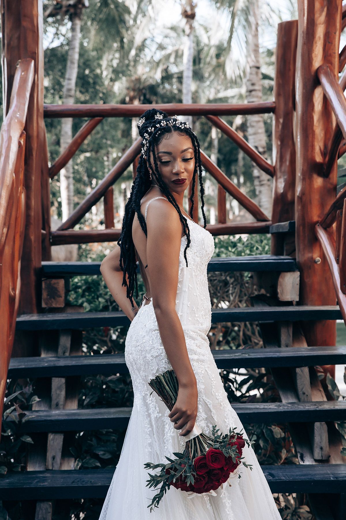 Bride holding a dark red and green bouquet while standing on rustic wooden deck surrounded by palm trees.