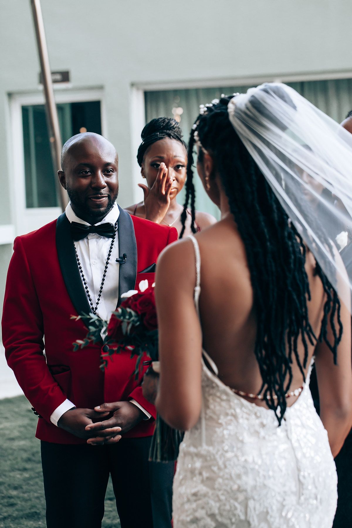 Emotional moment at a wedding ceremony with groom in red tuxedo and bride in white lace dress with long braids.