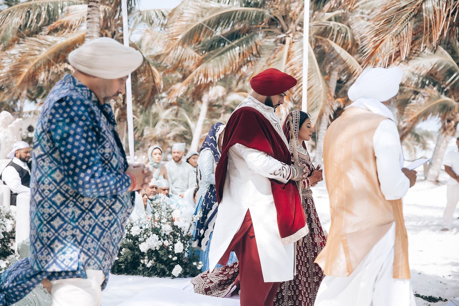 Wedding guests in traditional attire gather under palm trees at a beach ceremony.