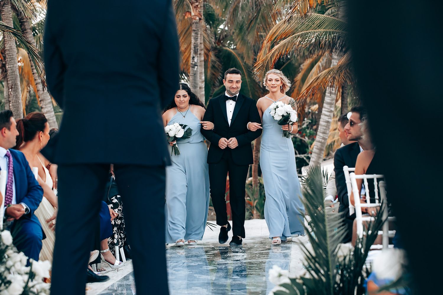 Elegant outdoor wedding ceremony with guests gathered under palm trees and white floral arrangements.