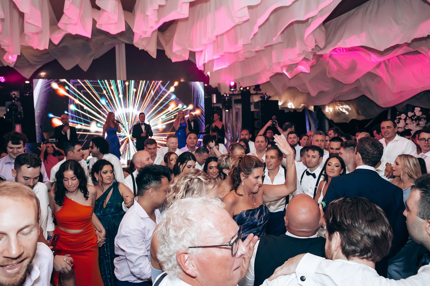 Crowded dance floor under pink lighting at an indoor wedding reception celebration.