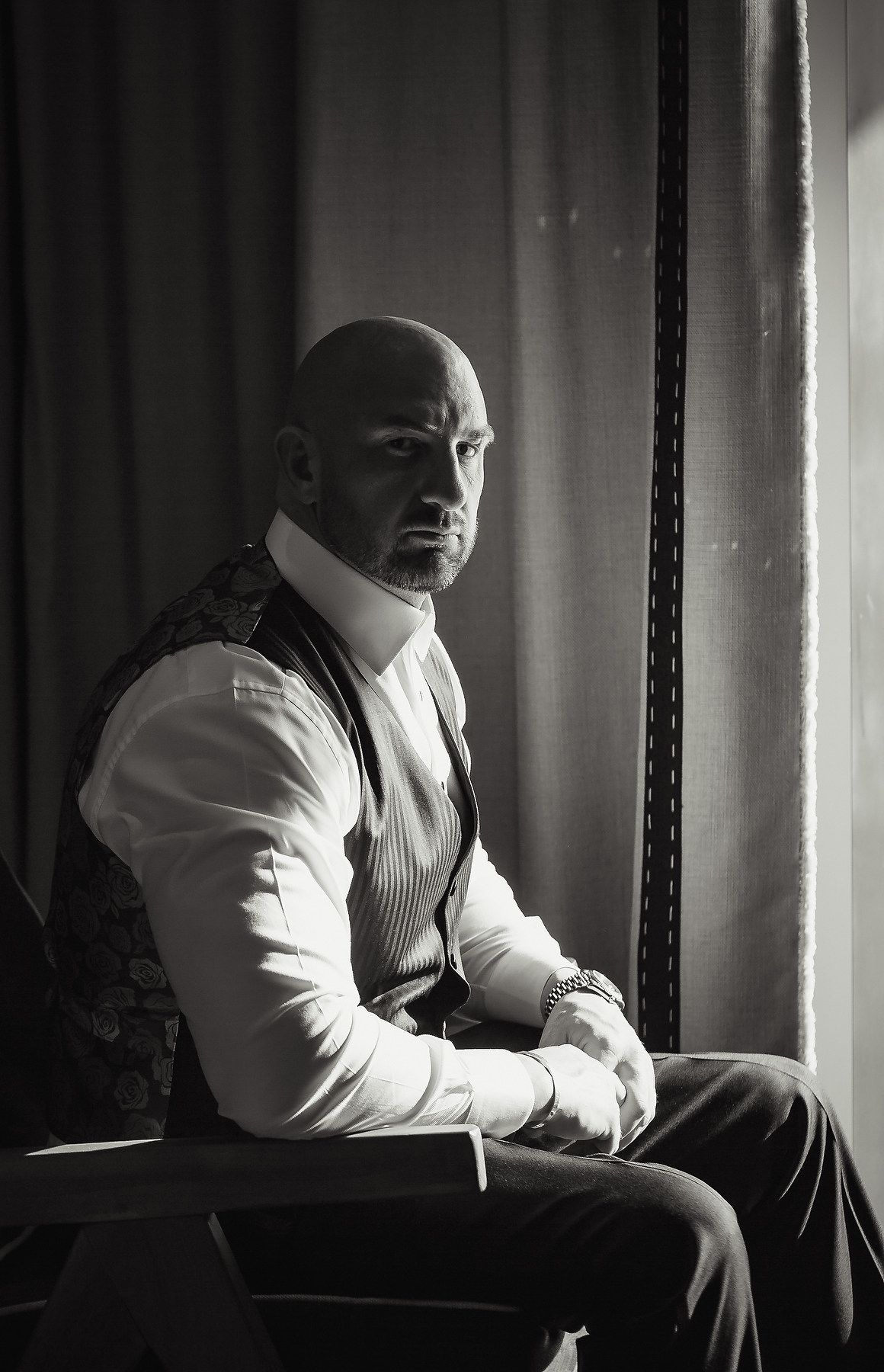 Dramatic black and white portrait of a groom in white shirt sitting by window light.
