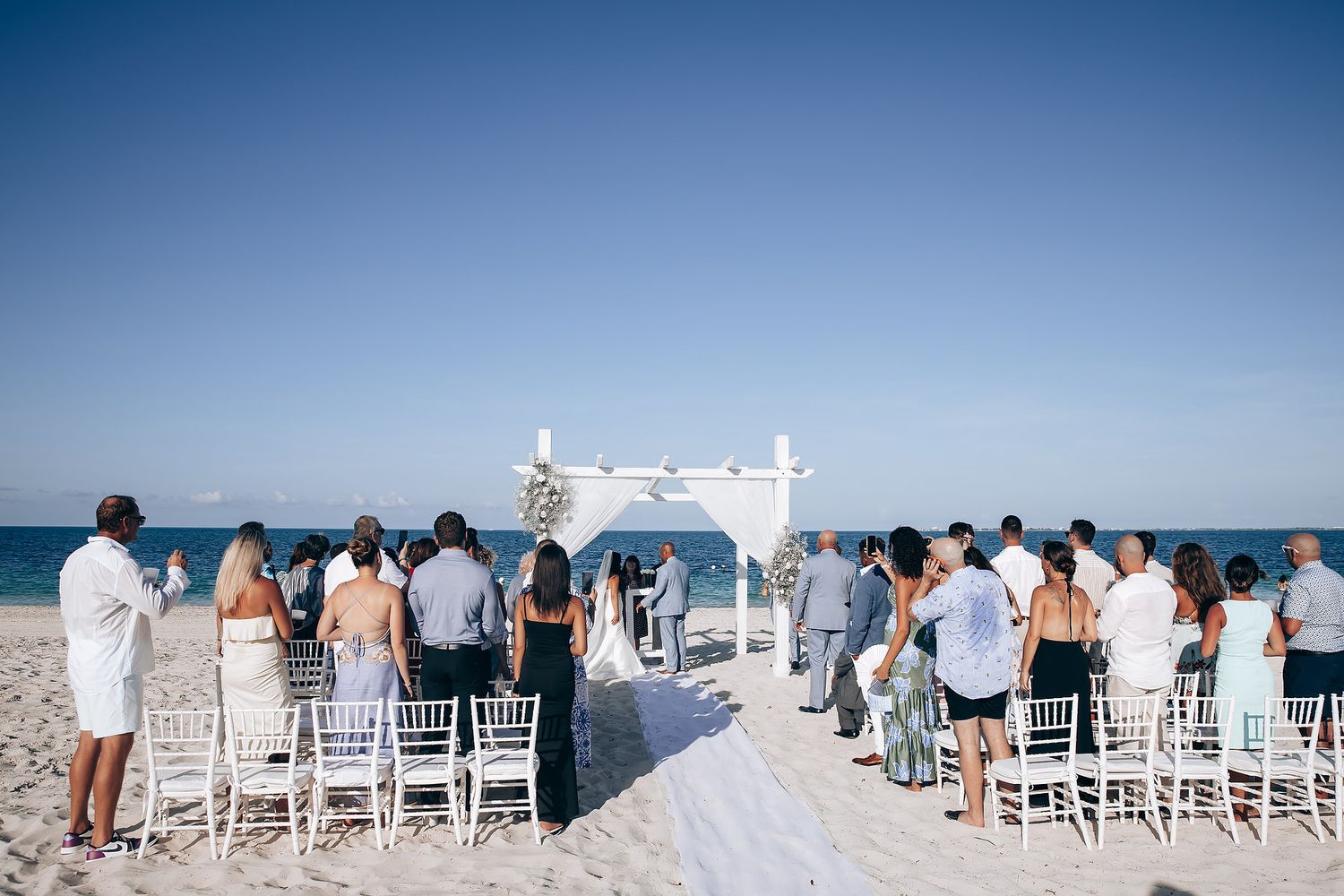 Beachfront wedding ceremony setup with white chairs against a bright blue ocean backdrop.