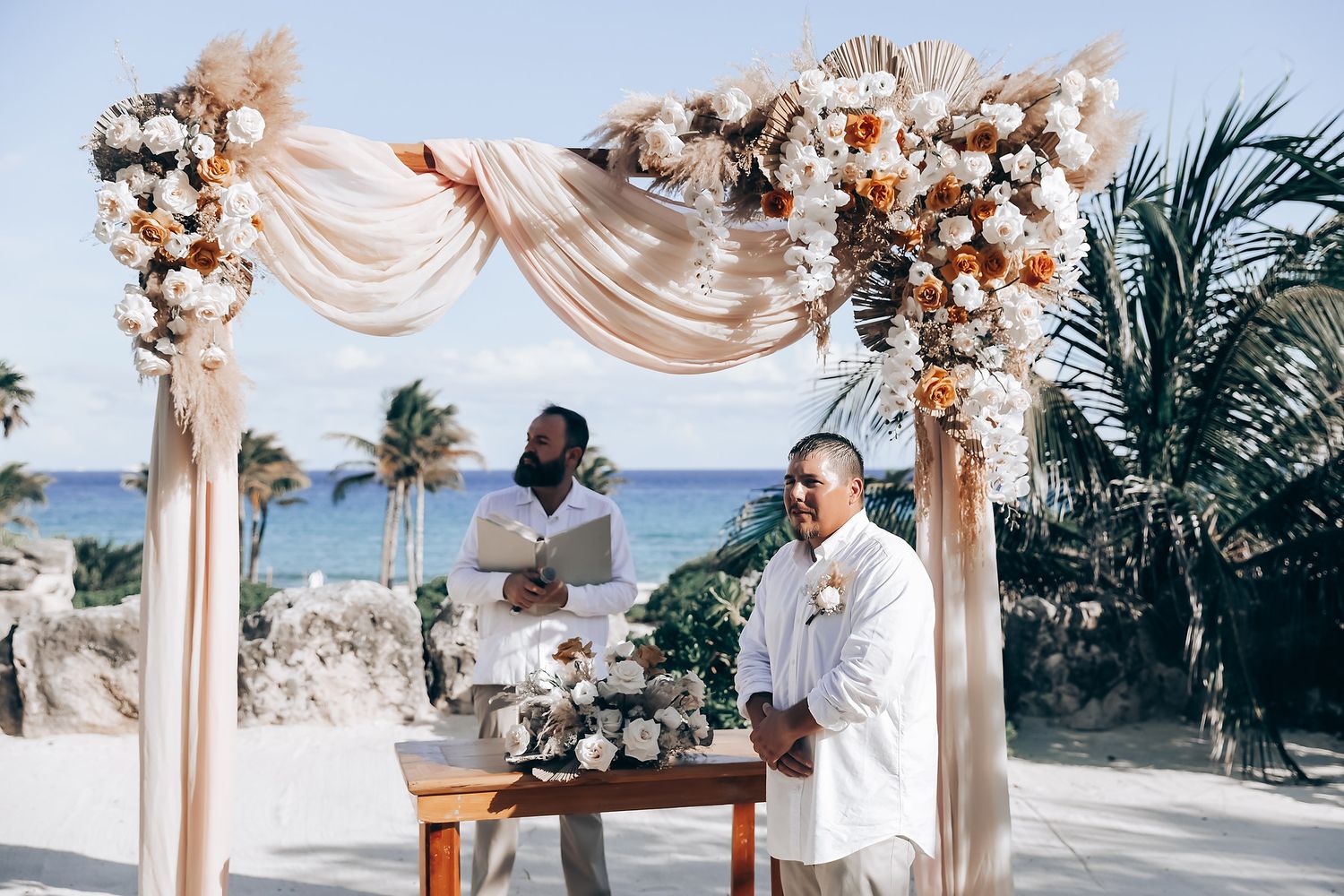 Elegant beach wedding ceremony setup with blush drapes and floral arrangements against an ocean backdrop.