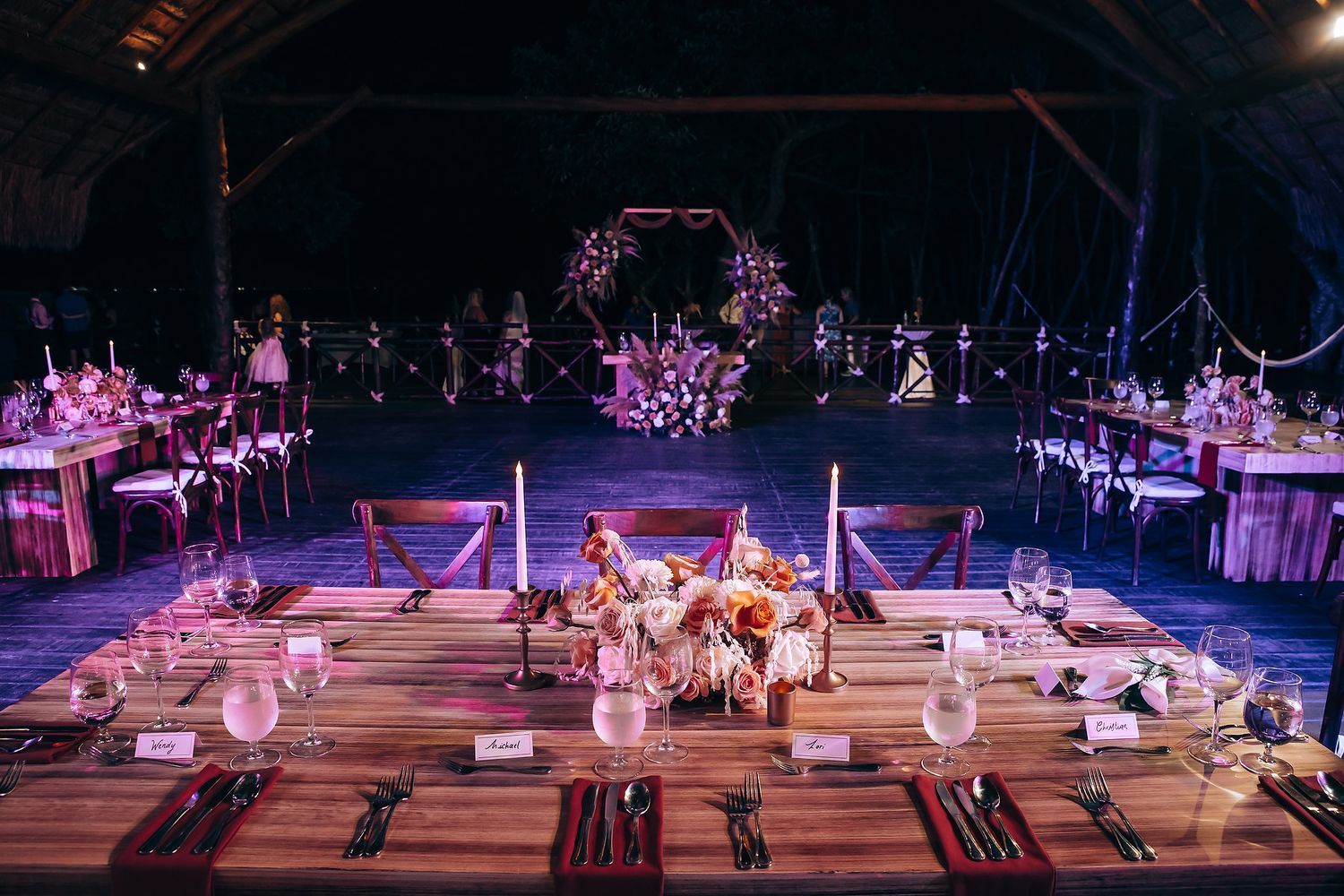 Outdoor wedding reception setup at night with illuminated hanging lanterns and rustic wooden tables.