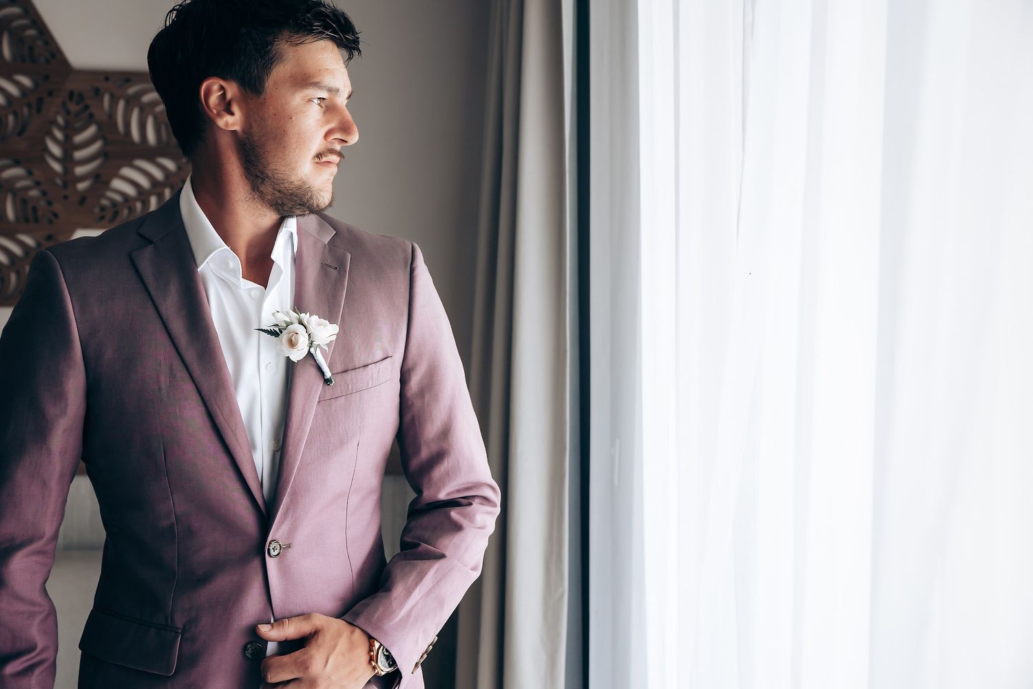 A person in a burgundy suit jacket stands thoughtfully by a window with natural light streaming in.