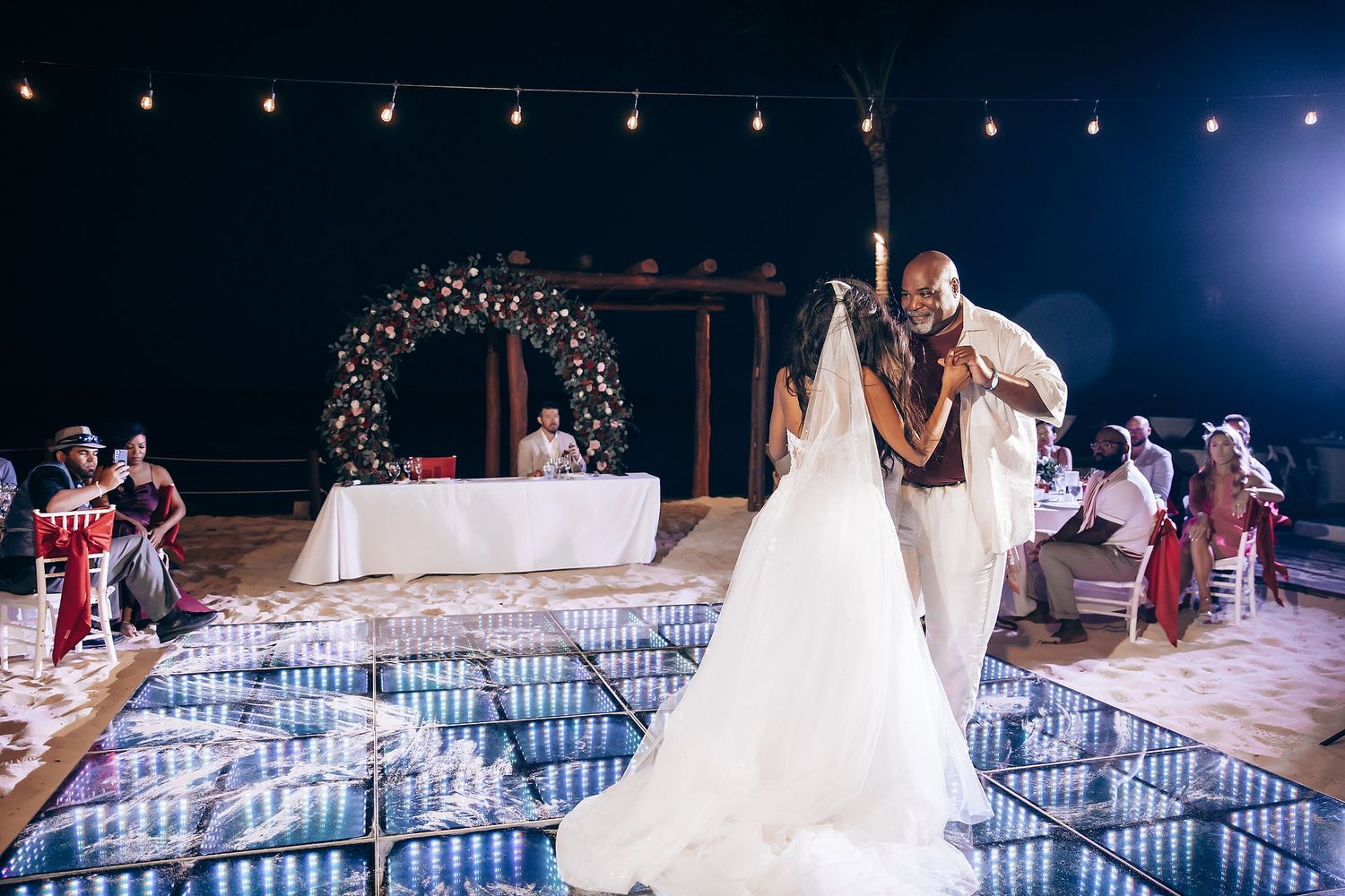 Elegant beach wedding reception setup with string lights and dance floor near the ocean.