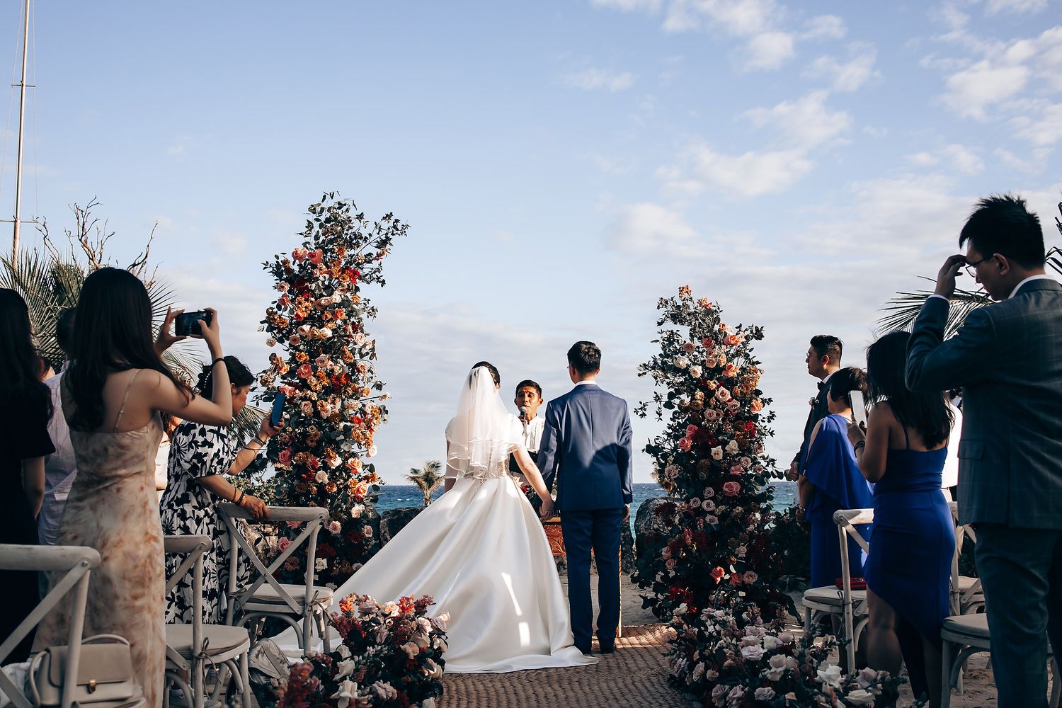 Wedding photographers capturing ceremony moments with floral arch and ocean backdrop.