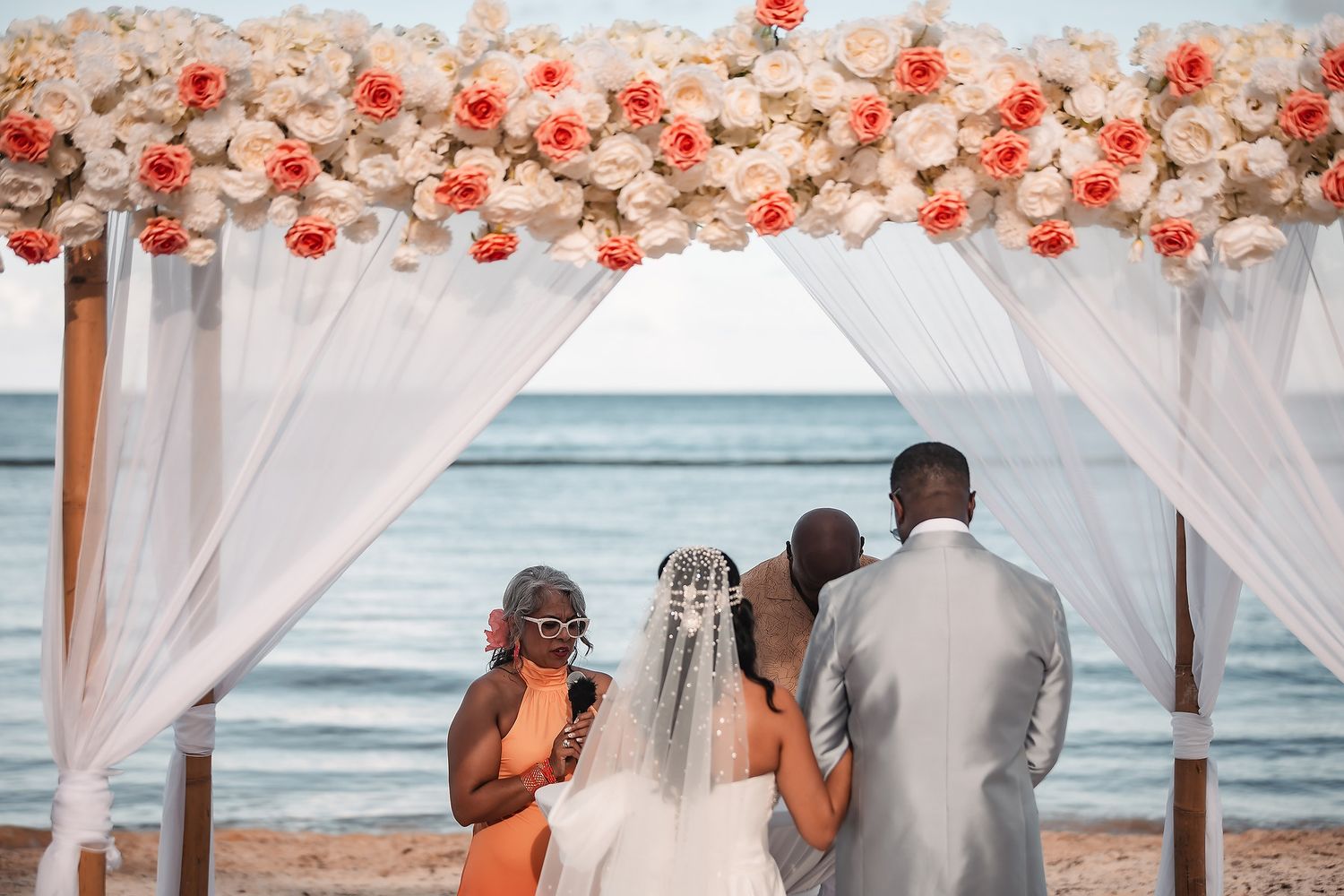 A beachfront wedding ceremony setup with coral floral arrangements and white draping overlooking the ocean.