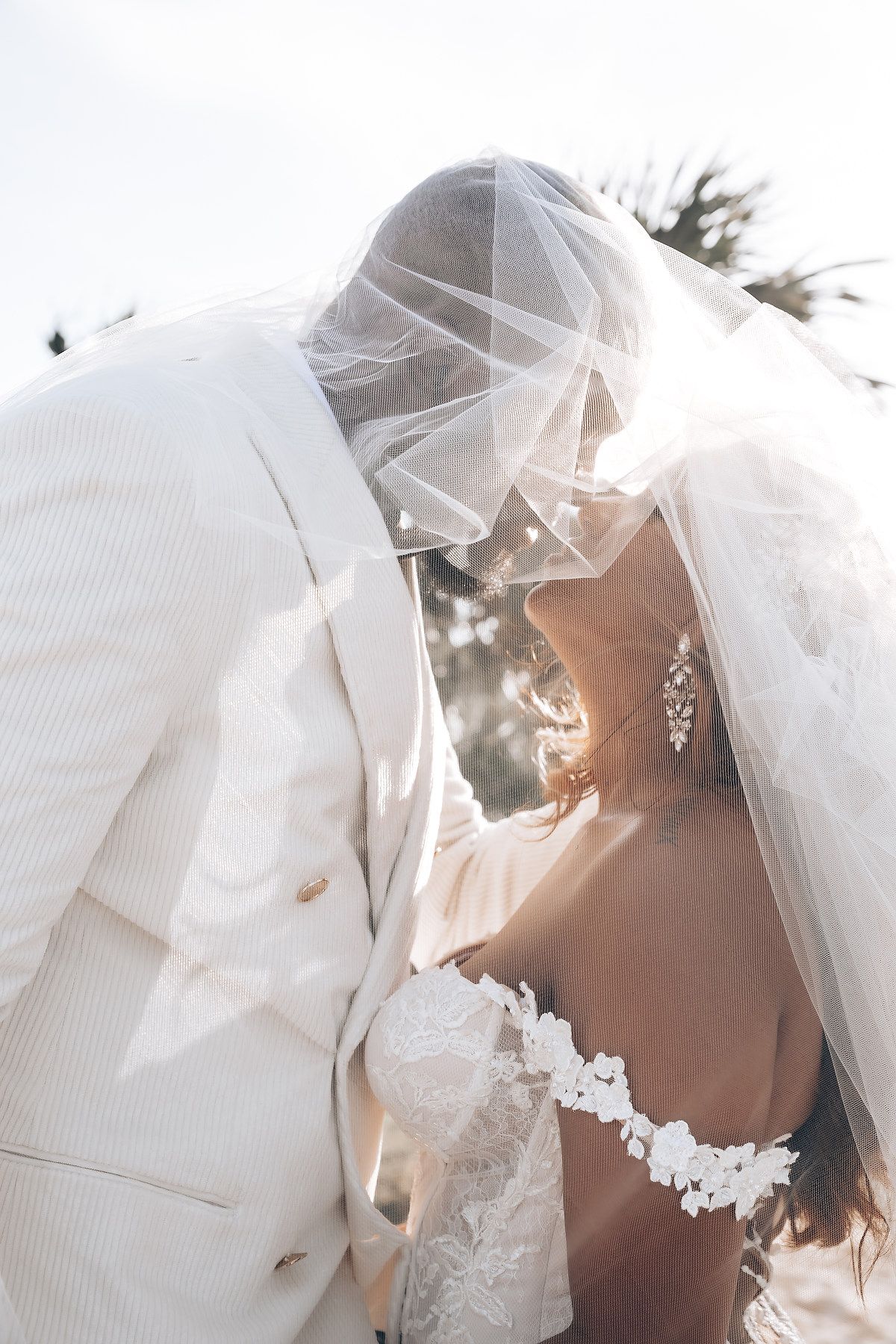 Romantic sunlit moment with bride in white dress and flowing veil against natural outdoor backdrop.