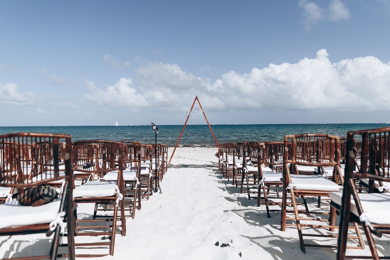 Wooden chairs arranged on a white sandy beach for a beach wedding ceremony with a triangular arch and ocean view.