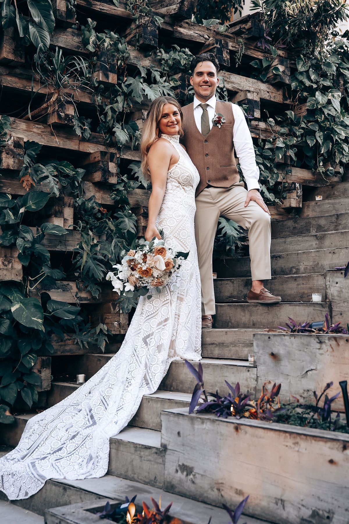 Wedding couple poses on rustic wooden steps surrounded by lush greenery and flowers.