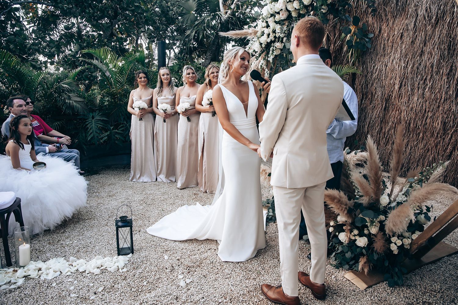 Wedding ceremony guests gathered under palm trees with elegant white and beige decor.