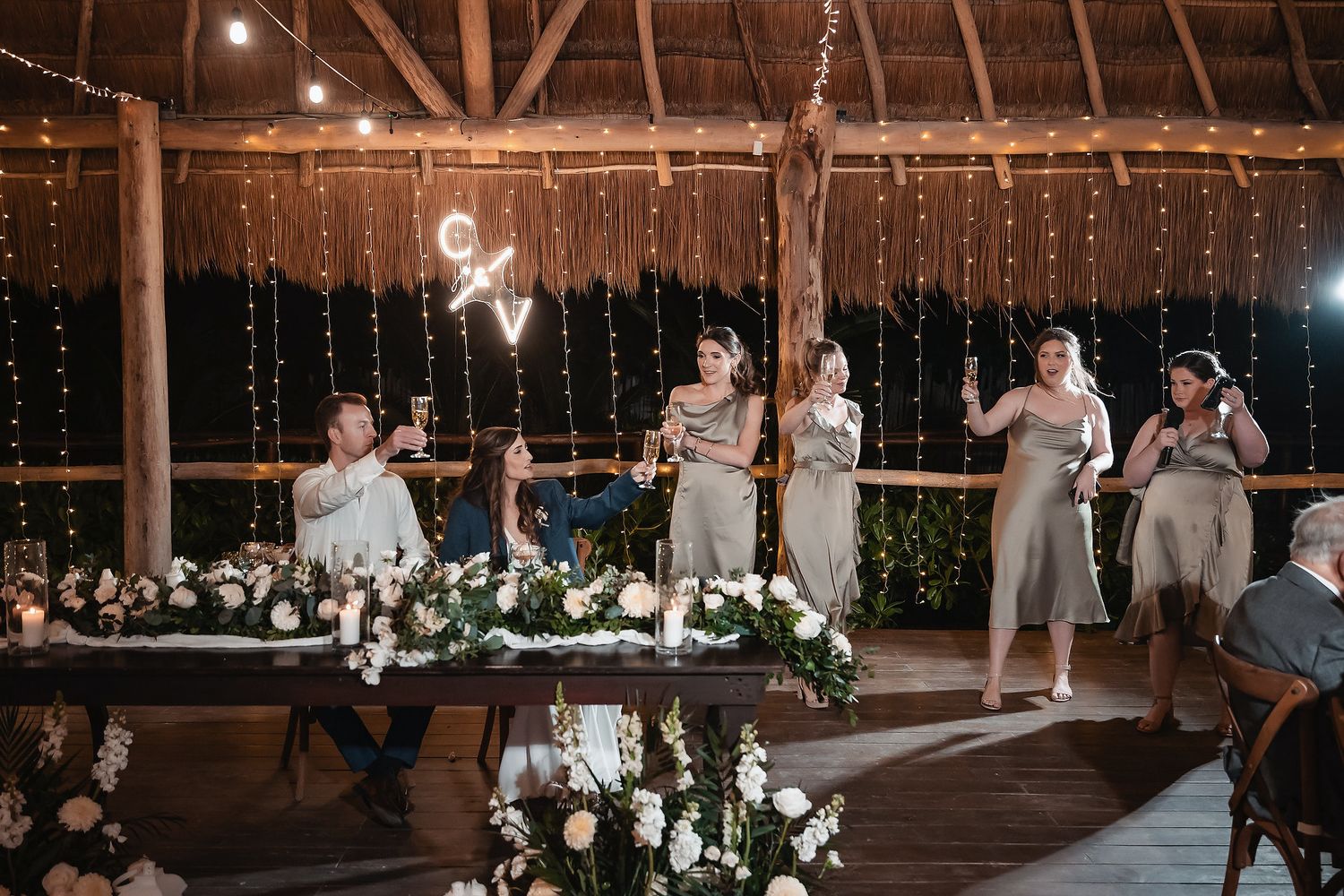 Bridesmaids giving toasts at elegant outdoor wedding reception with fairy lights and white floral arrangements.