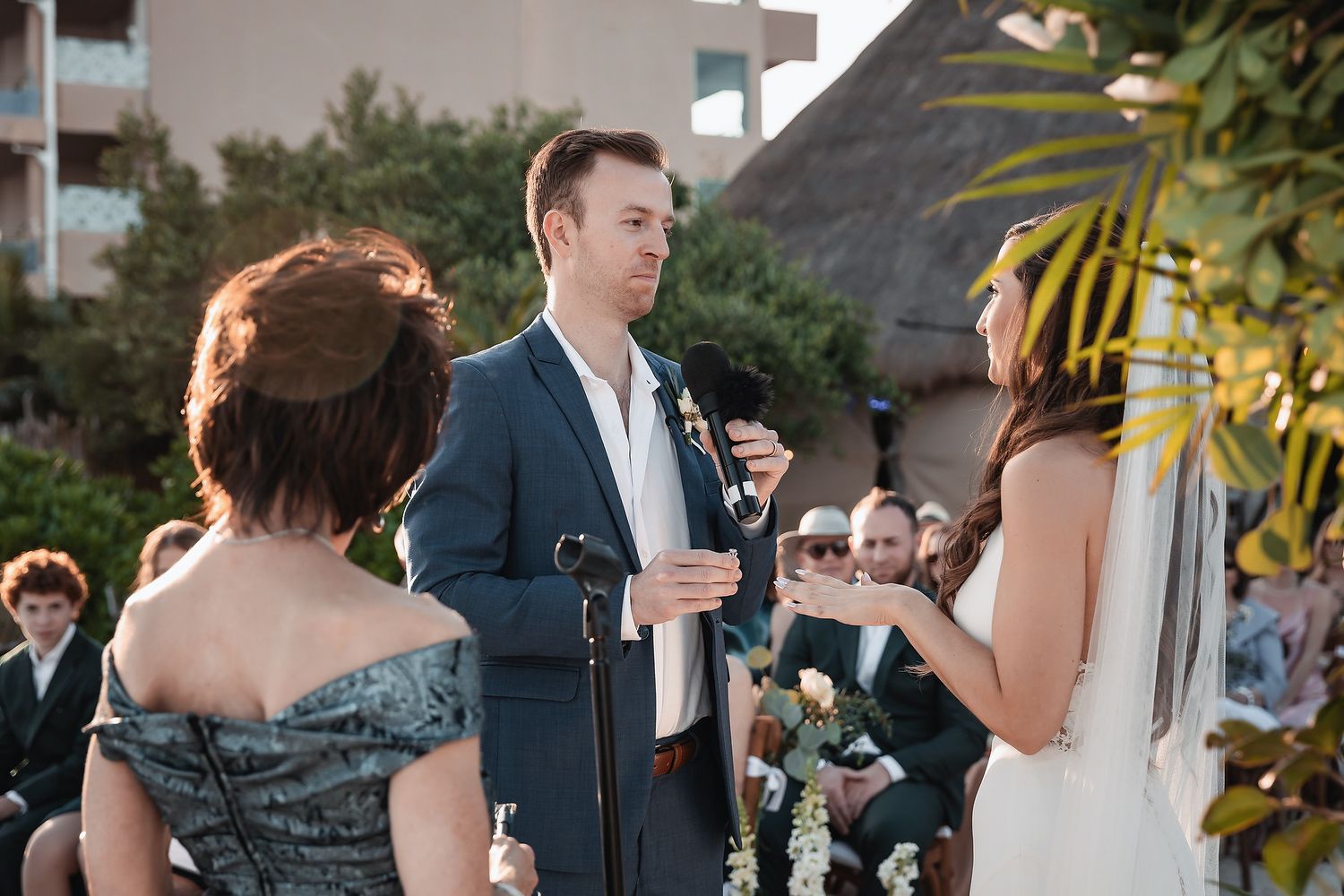 Groom in navy suit exchanges vows with bride at outdoor wedding ceremony with guests seated behind.