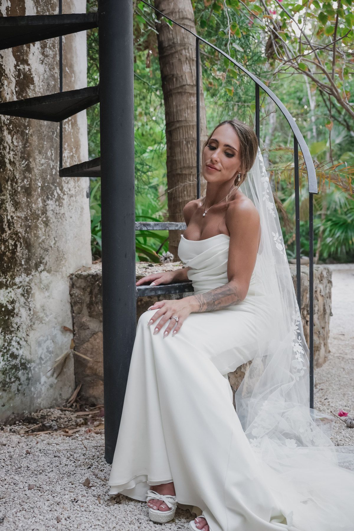 Bride in white strapless gown and veil sits gracefully by spiral staircase surrounded by lush greenery.