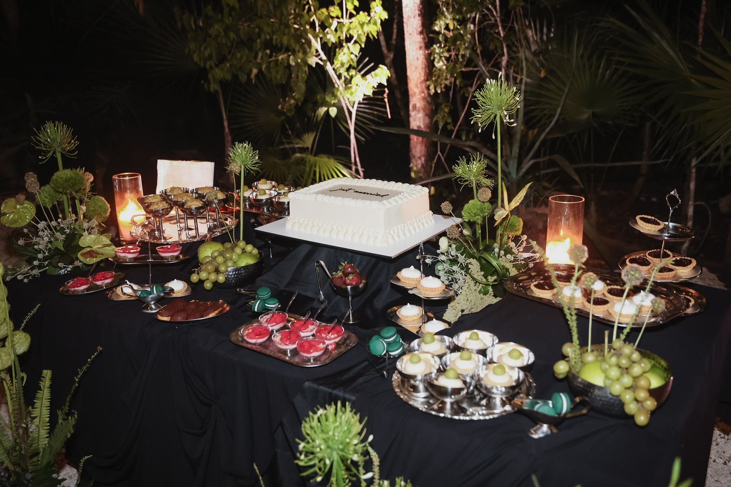 Elegant outdoor dessert table with white cake, candles, and assorted treats on black tablecloth at night.