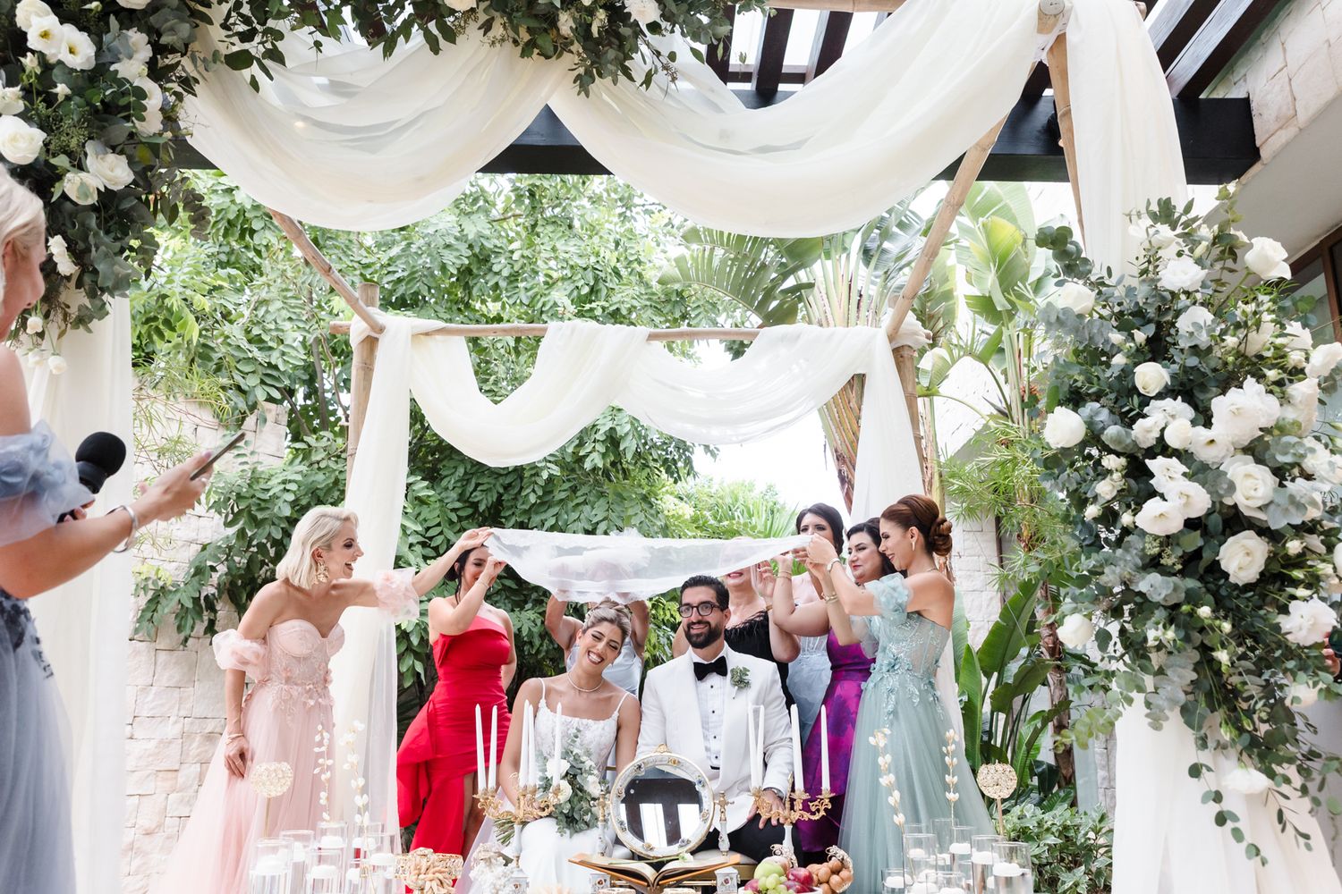 Elegant outdoor wedding ceremony with bride and groom under floral arch, surrounded by bridesmaids in colorful dresses.