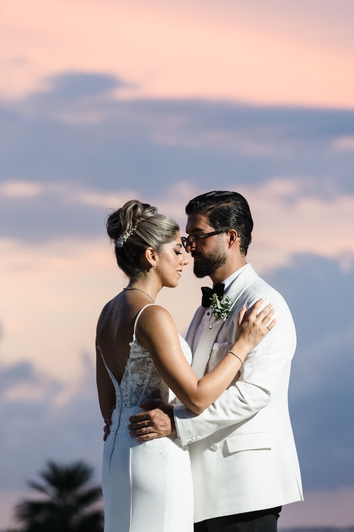 Bride and groom share a romantic embrace at sunset, with pink and blue clouds in the background.