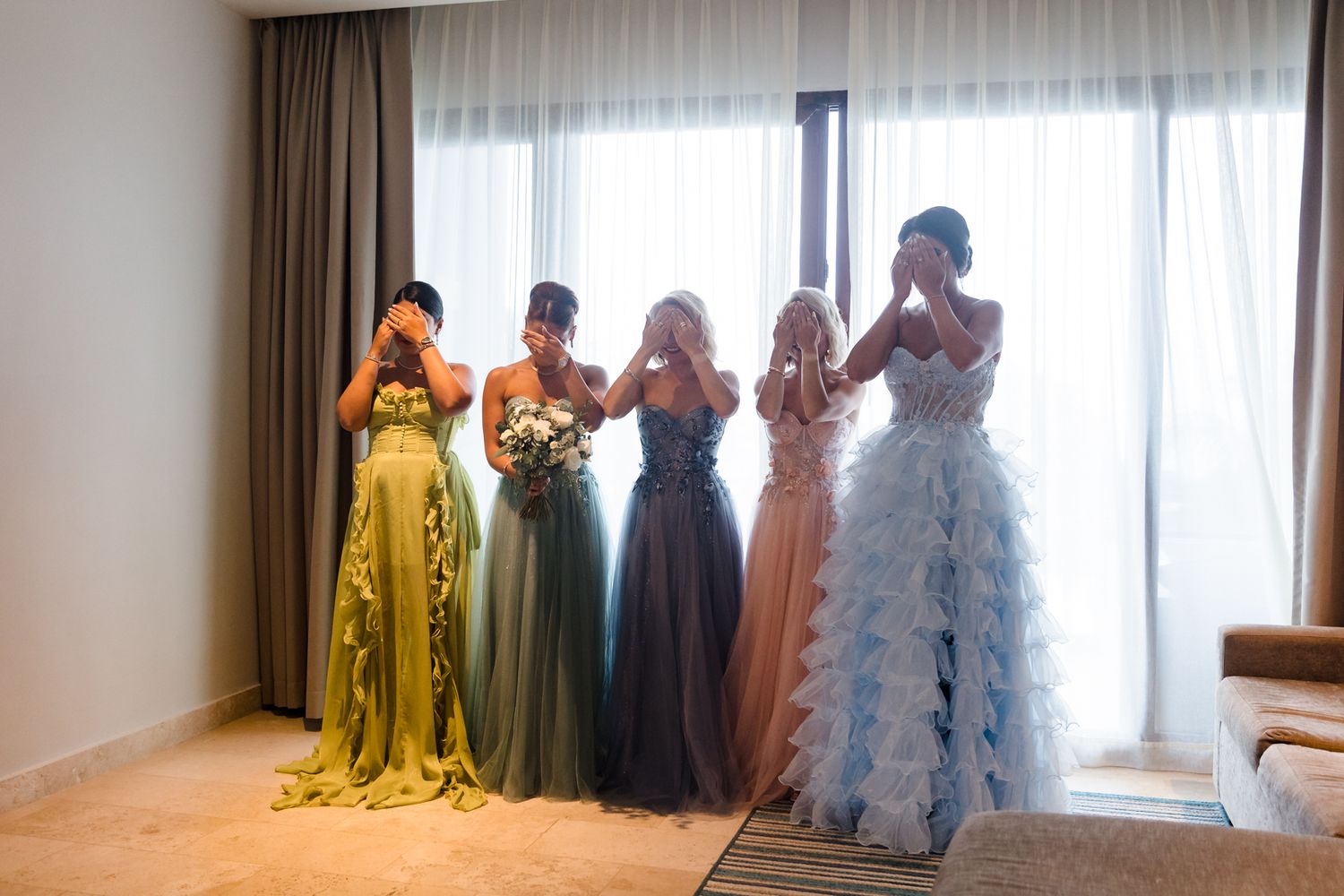 Five bridesmaids in colorful gowns covering their faces with emotion near a bright window in an elegant hotel room.