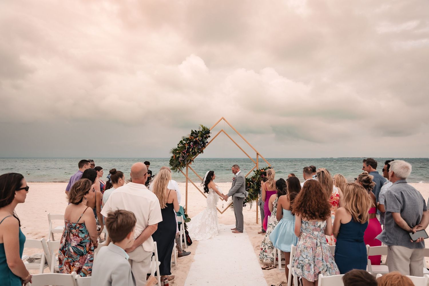 Wedding guests gather on a sandy beach with an overcast sky in the background.