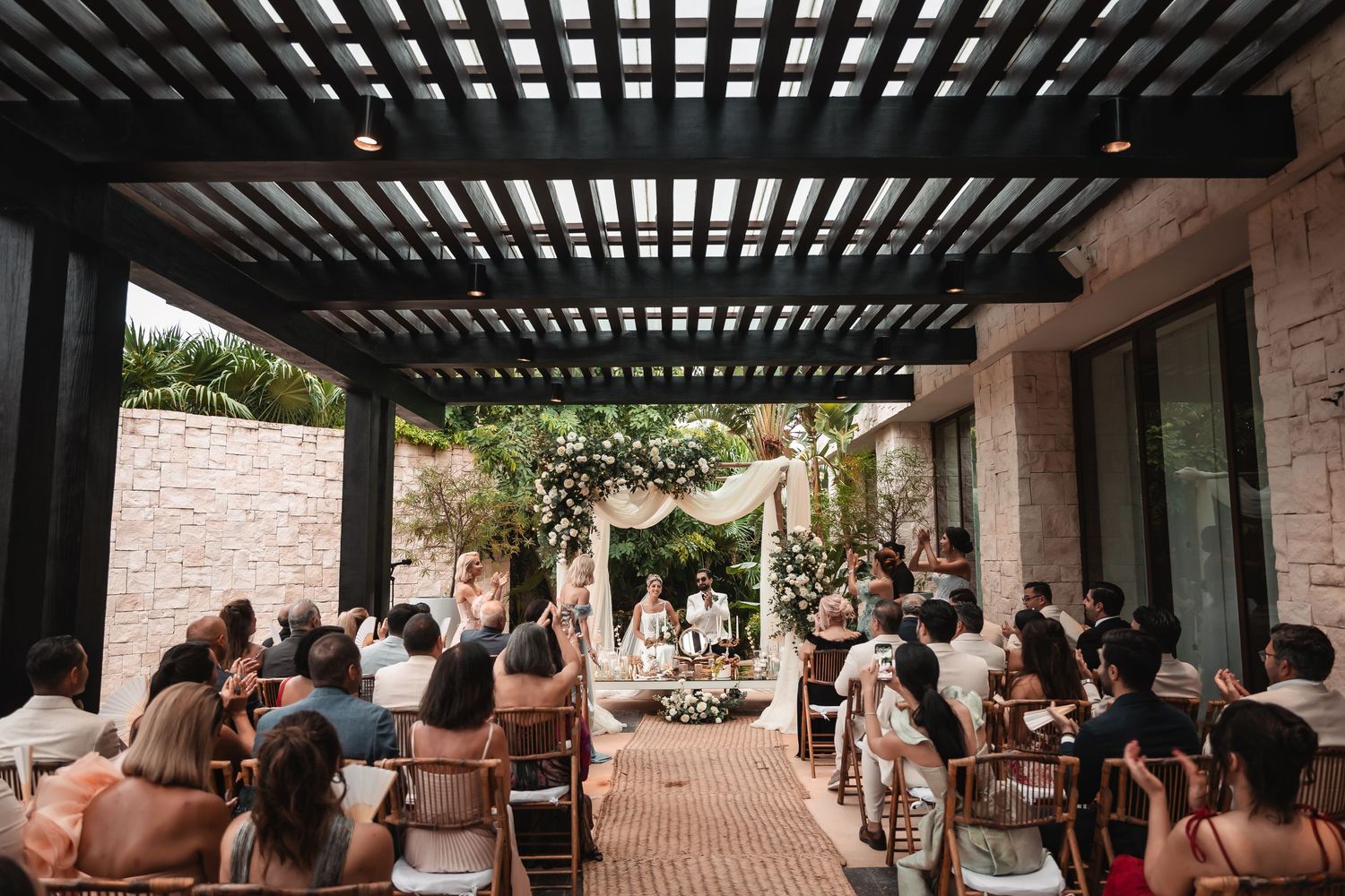 Wide view of covered outdoor wedding reception space with wooden pergola and stone architecture.