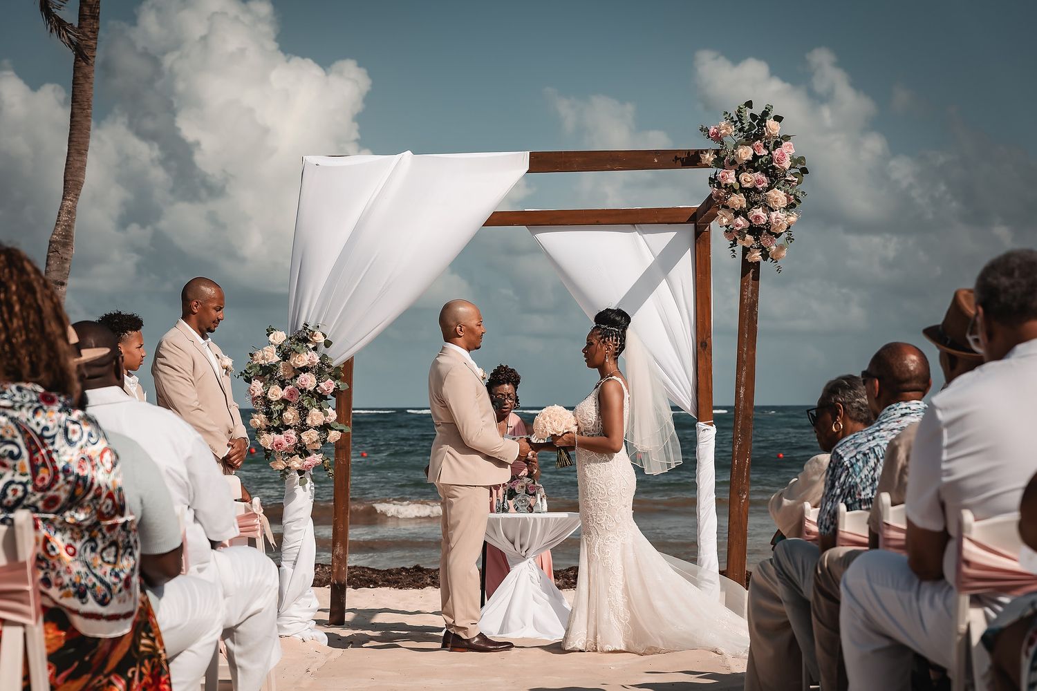 Wedding ceremony in progress at a beachfront location with white decor and floral arrangements.