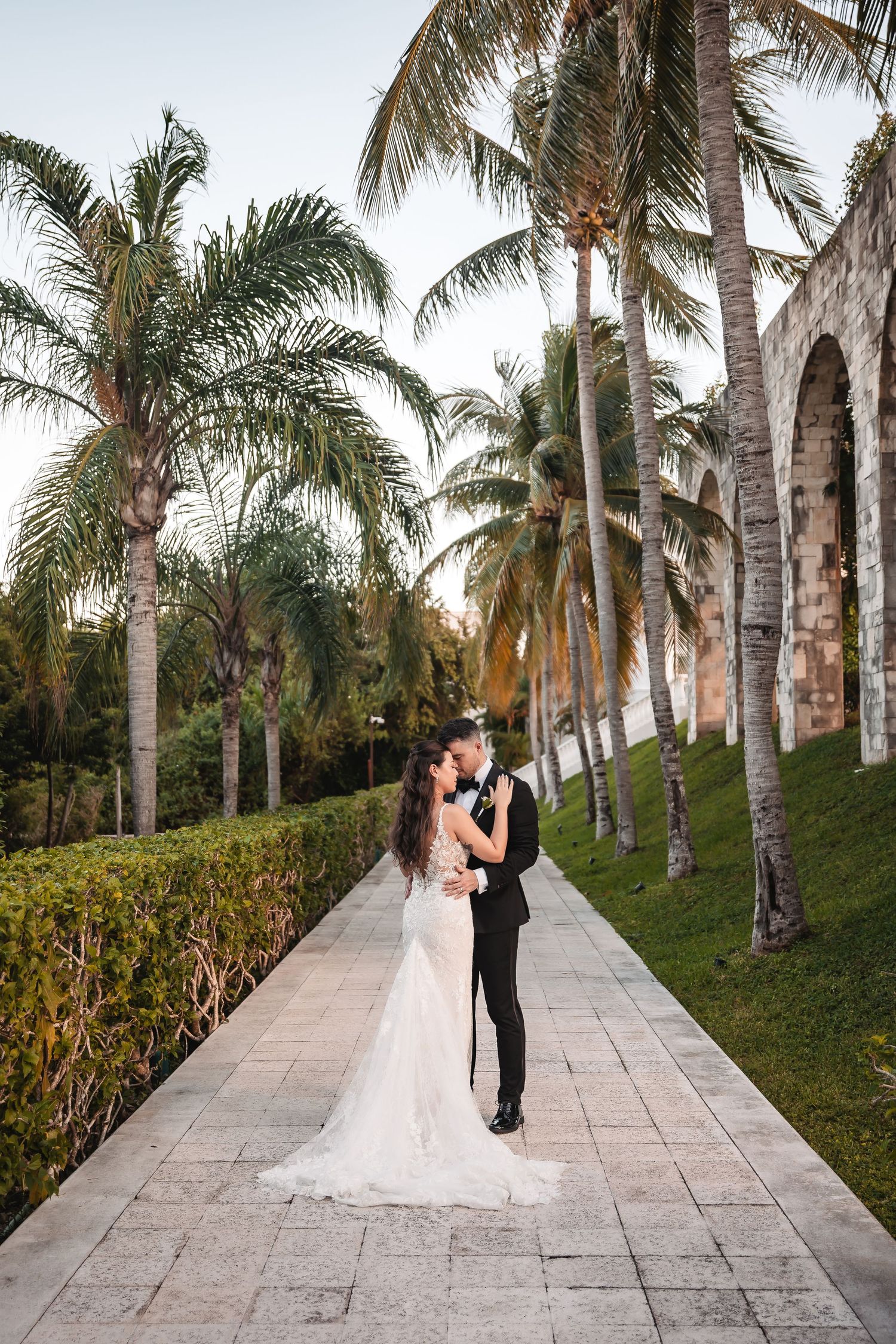 Wedding couple shares a romantic moment on a stone pathway lined with majestic palm trees.
