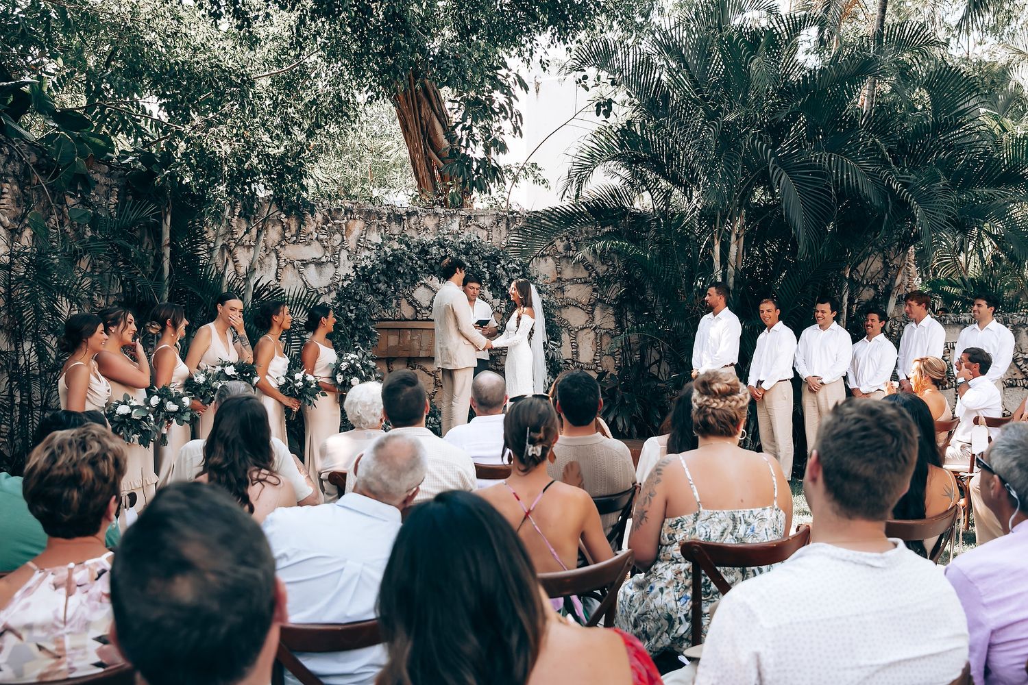 Wedding guests in white and light-colored attire gather outdoors under trees.
