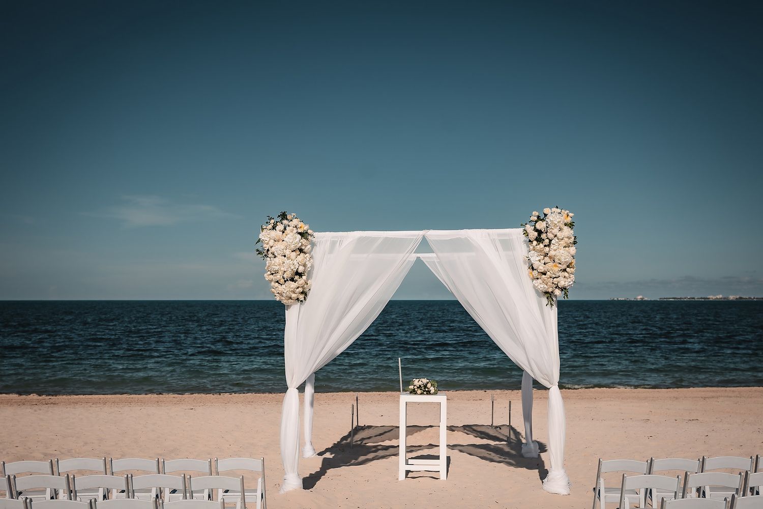 White fabric wedding arch with floral arrangements stands elegantly on a beach against blue ocean waters.