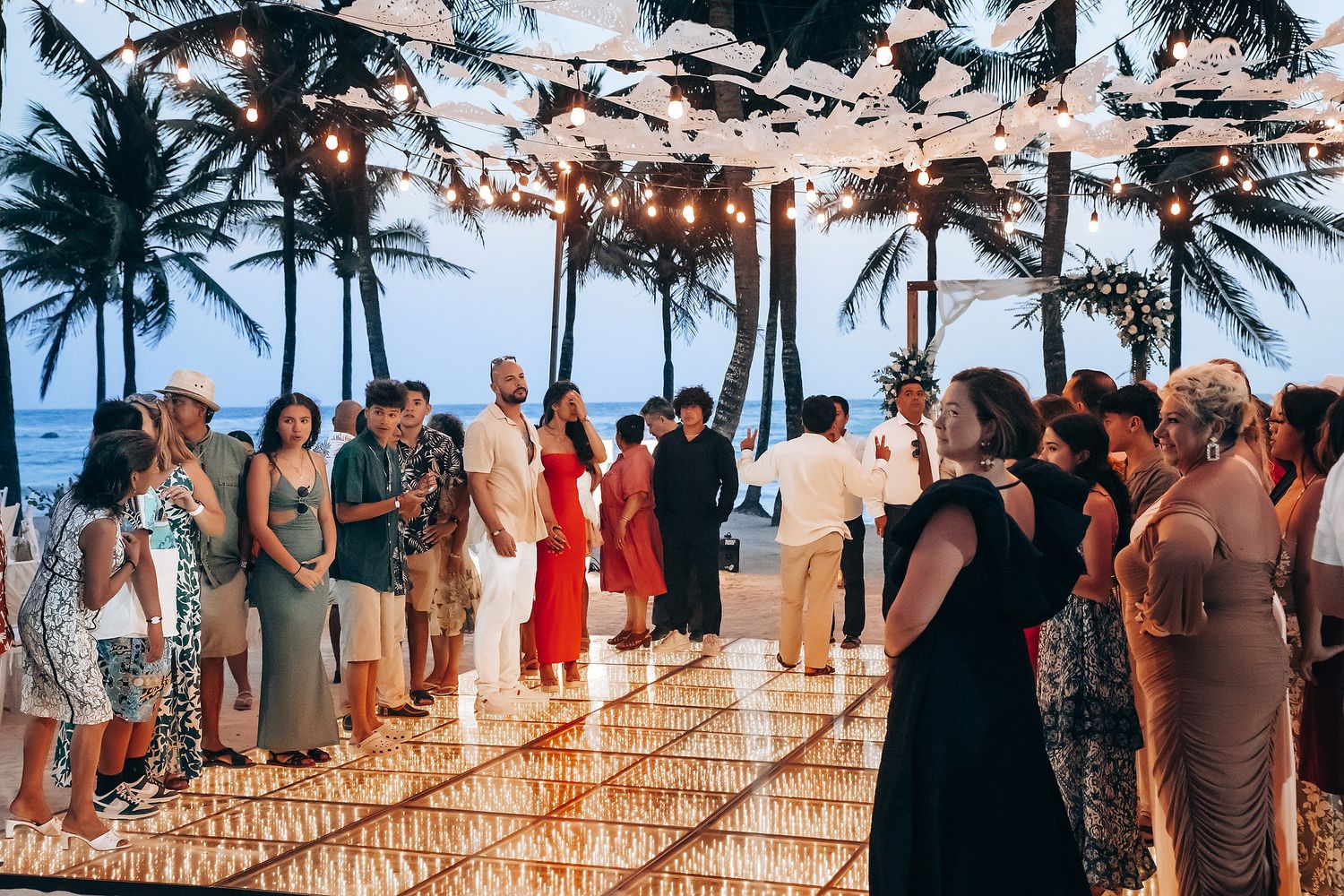 Wedding guests mingle at a beachfront reception with string lights and palm trees.