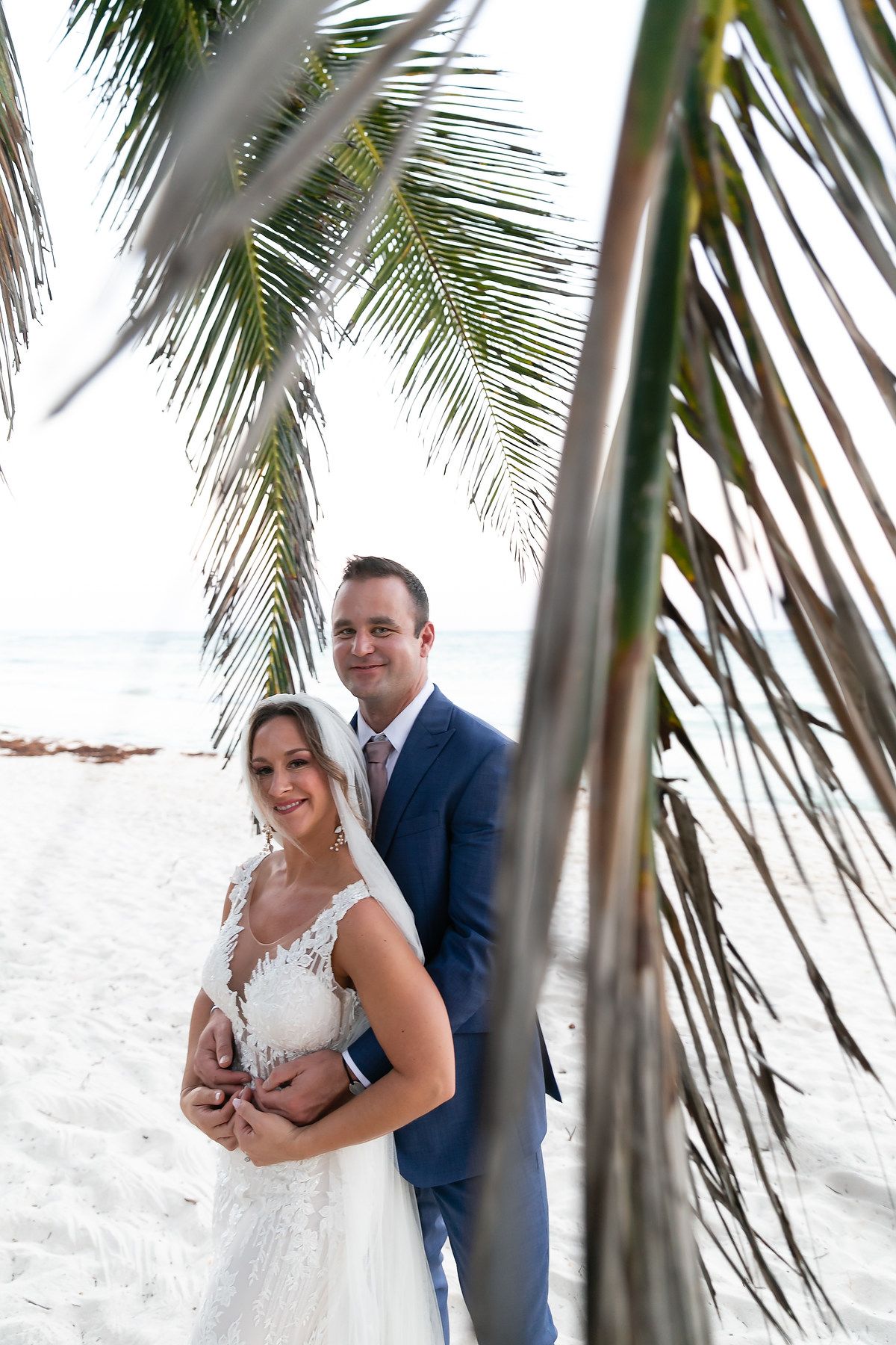 A romantic beach wedding photo framed by palm fronds with white sand and tropical scenery in the background.