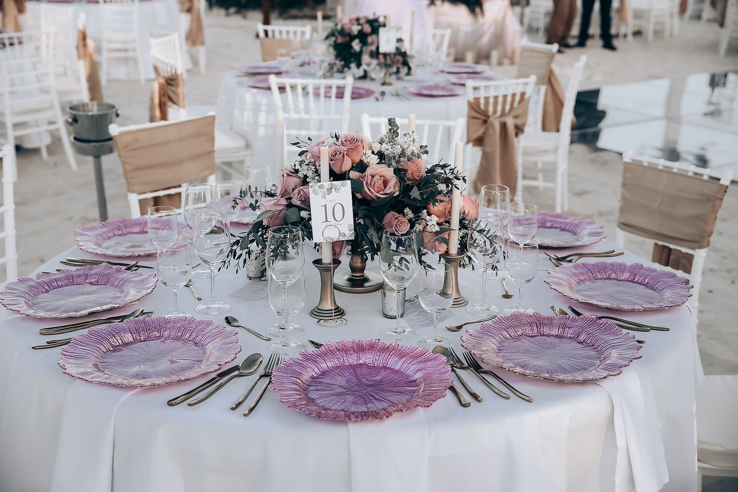 Elegant wedding reception tables set with purple charger plates and floral centerpieces in a light-filled venue.